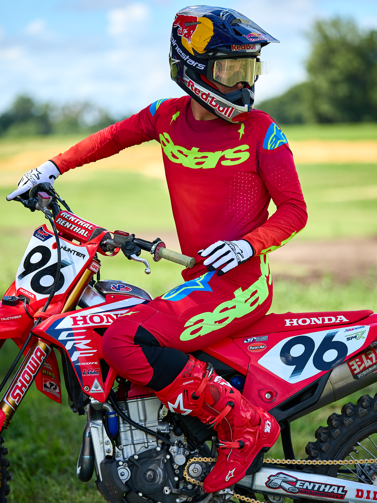 Motocross rider on a red Honda motorcycle wearing the Alpinestars 2025 Supertech Maker Gear Set in Red, featuring a red jersey with neon green 'astars' branding and blue shoulder accents, matching red pants, solid red boots, white gloves, and a Red Bull helmet, professional racing kit for off-road performance