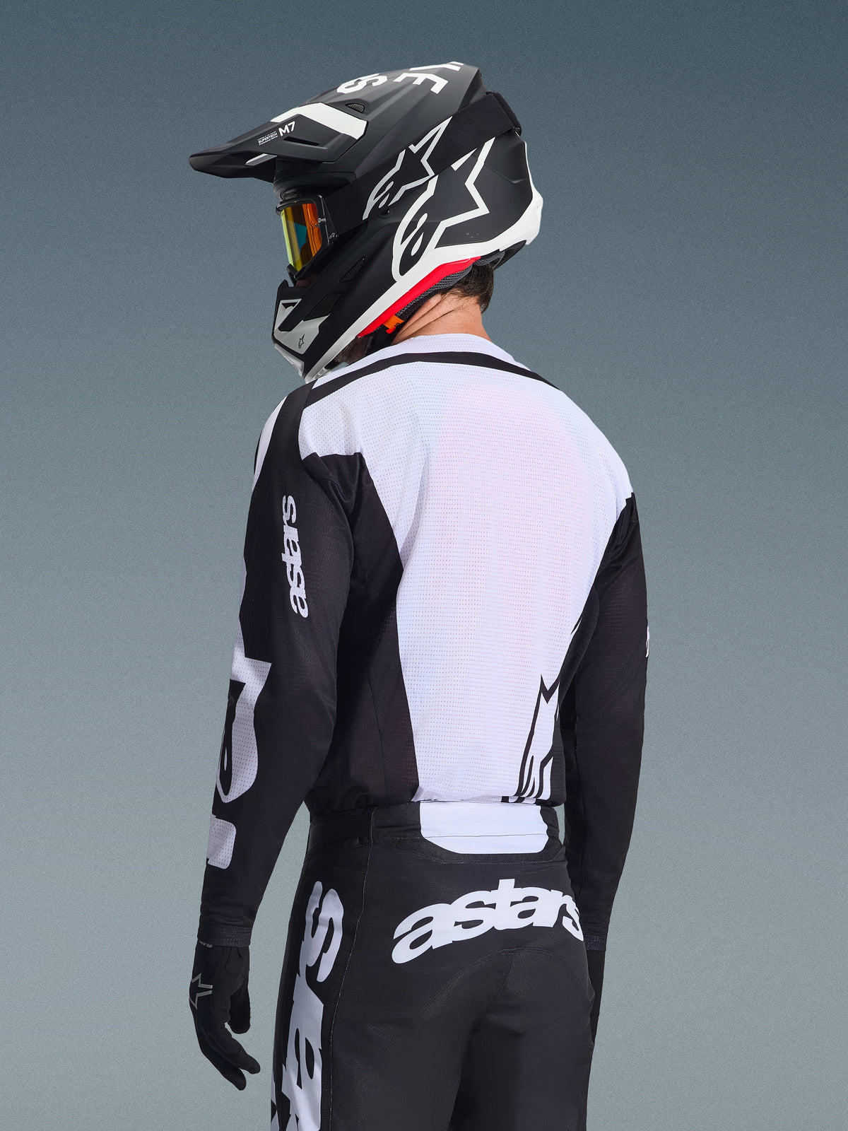 Motocyklista motocrossowy widziany od tyłu w koszulce Alpinestars 2026 Racer Air Riway, czarno-biała, z białym oddychającym panelem z siateczki na plecach i czarnymi rękawami z brandingiem „astars”, pasujące czarne spodnie z białym logo, czarny kask z białymi akcentami, profesjonalny sprzęt off-road o wysokiej wydajności w wysokich temperaturach.