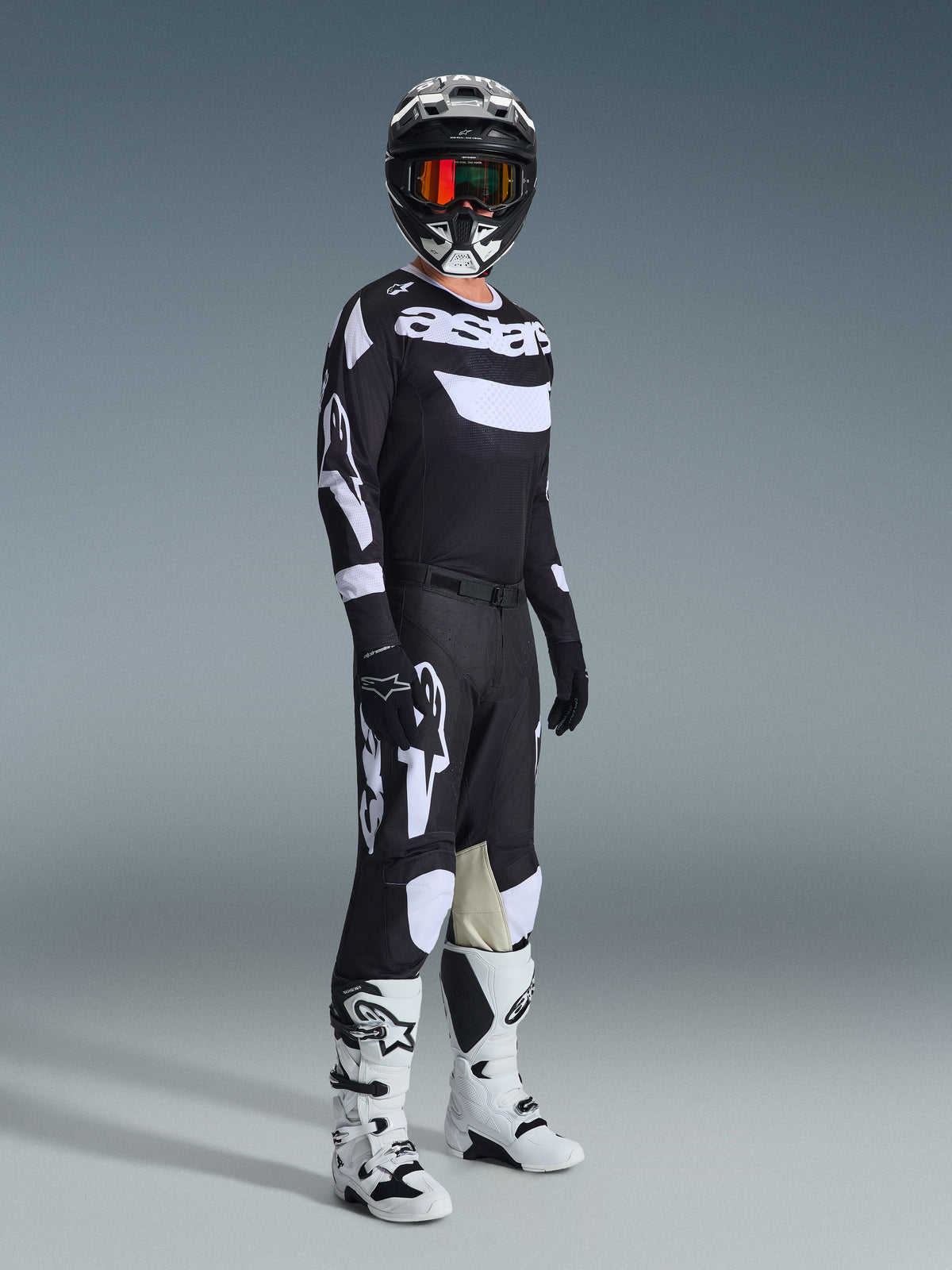 Rider motocrossowy w pełnym stroju wyścigowym, koszulka 2026 Racer Air Riway w kolorze czarno-białym, pasujące czarne spodnie z dużymi białymi logo Alpinestars, czarny kask z białymi akcentami i pomarańczowymi goglami, biało-czarne buty, profesjonalny zestaw do jazdy off-road