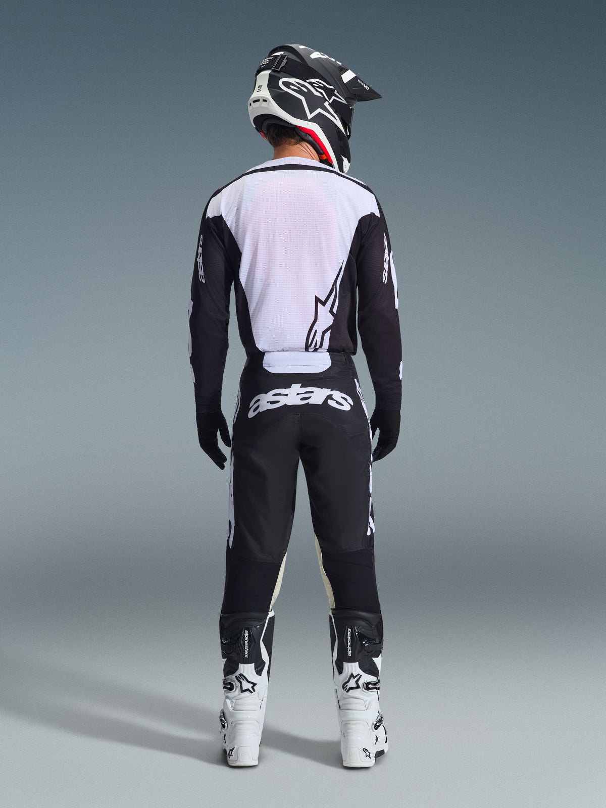 Widok z tyłu motocyklisty motocrossowego w koszulce Alpinestars 2026 Racer Air Riway w kolorze czarno-białym, z oddychającym panelem z siatki na plecach, czarnymi spodniami z białym logo „astars”, czarno-białym kaskiem i białymi butami wyścigowymi.