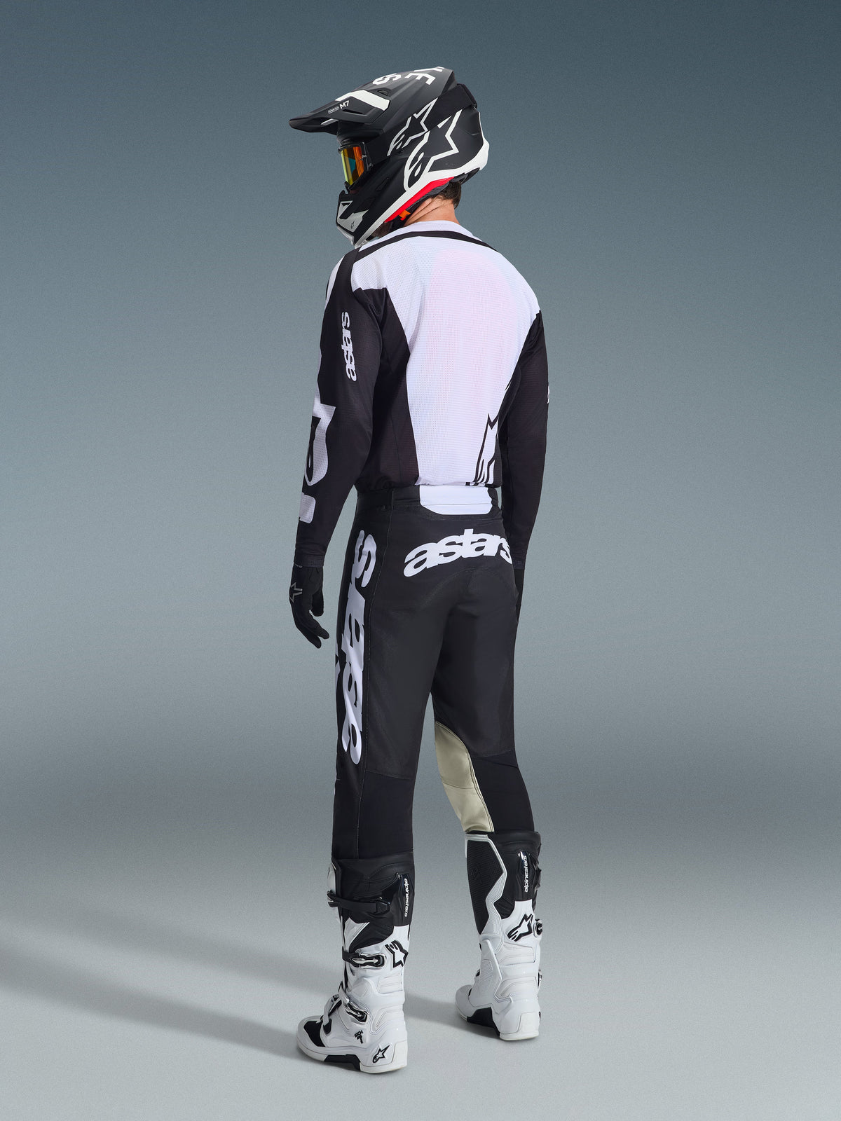 Motocyklista motocrossowy widoczny z tyłu, koszulka Alpinestars 2026 Racer Air Riway w kolorze czarno-białym, duży biały panel z siatki na plecach zapewniający przepływ powietrza, czarne spodnie, czarno-biały kask i białe buty wyścigowe, profesjonalny zestaw odzieży off-road