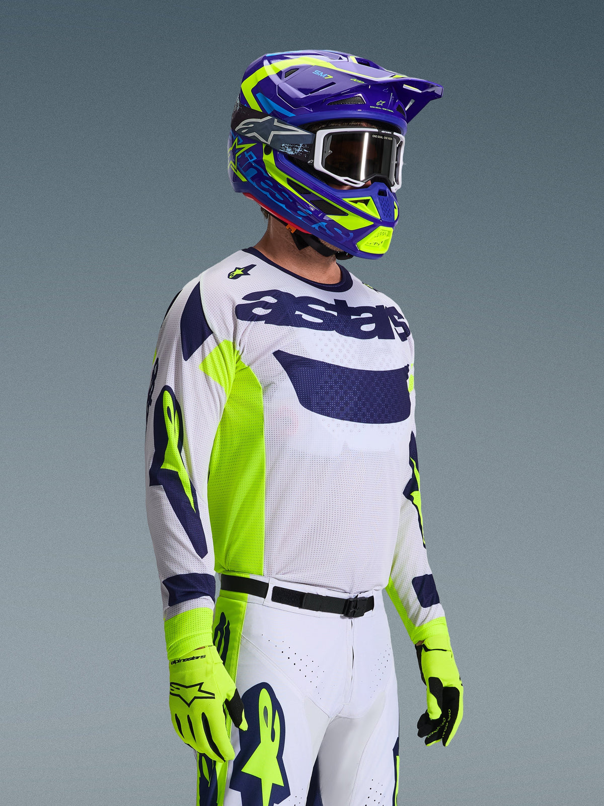 Motocyklista motocrossowy w koszulce Alpinestars 2026 Racer Air Riway, jasnoszara z akcentami w kolorze żółtym fluo i niebieskim, w zestawie z dopasowanym niebiesko-żółtym kaskiem i jaskrawożółtymi rękawicami, oddychająca konstrukcja z siateczki, profesjonalna stylistyka wyścigowa do jazdy off-road.