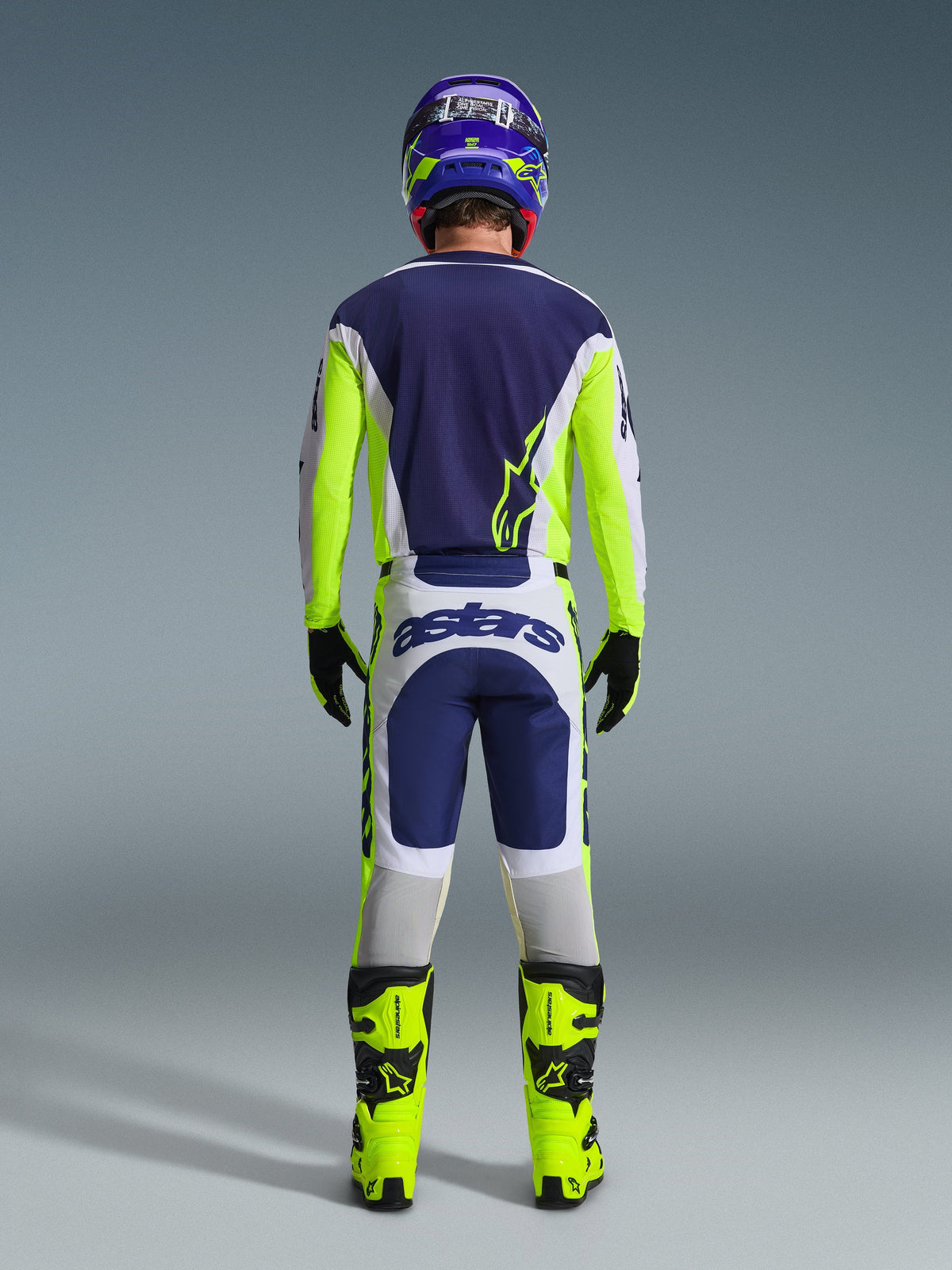 Widok z tyłu zawodnika motocrossowego w koszulce Alpinestars 2026 Racer Air Riway i pasujących spodniach w kolorze jasnoszarym, żółtym fluo i niebieskim, z jaskrawożółtymi butami fluo i dopasowanym kaskiem, prezentujący kompletny zestaw odzieży wyścigowej off-road.