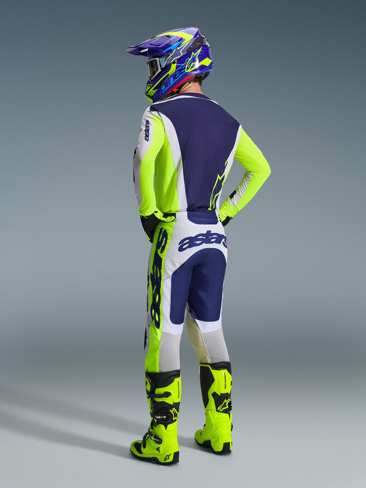 Zawodnik motocrossowy widoczny od tyłu w koszulce 2026 Racer Air Riway Jersey w kolorze jasnoszarym, fluorescencyjnym żółtym i niebieskim, pasujące spodnie z wyraźnym brandingiem, jaskrawożółte buty motocrossowe fluo oraz niebiesko-żółty kask, kompletny zestaw odzieży wyścigowej off-road.