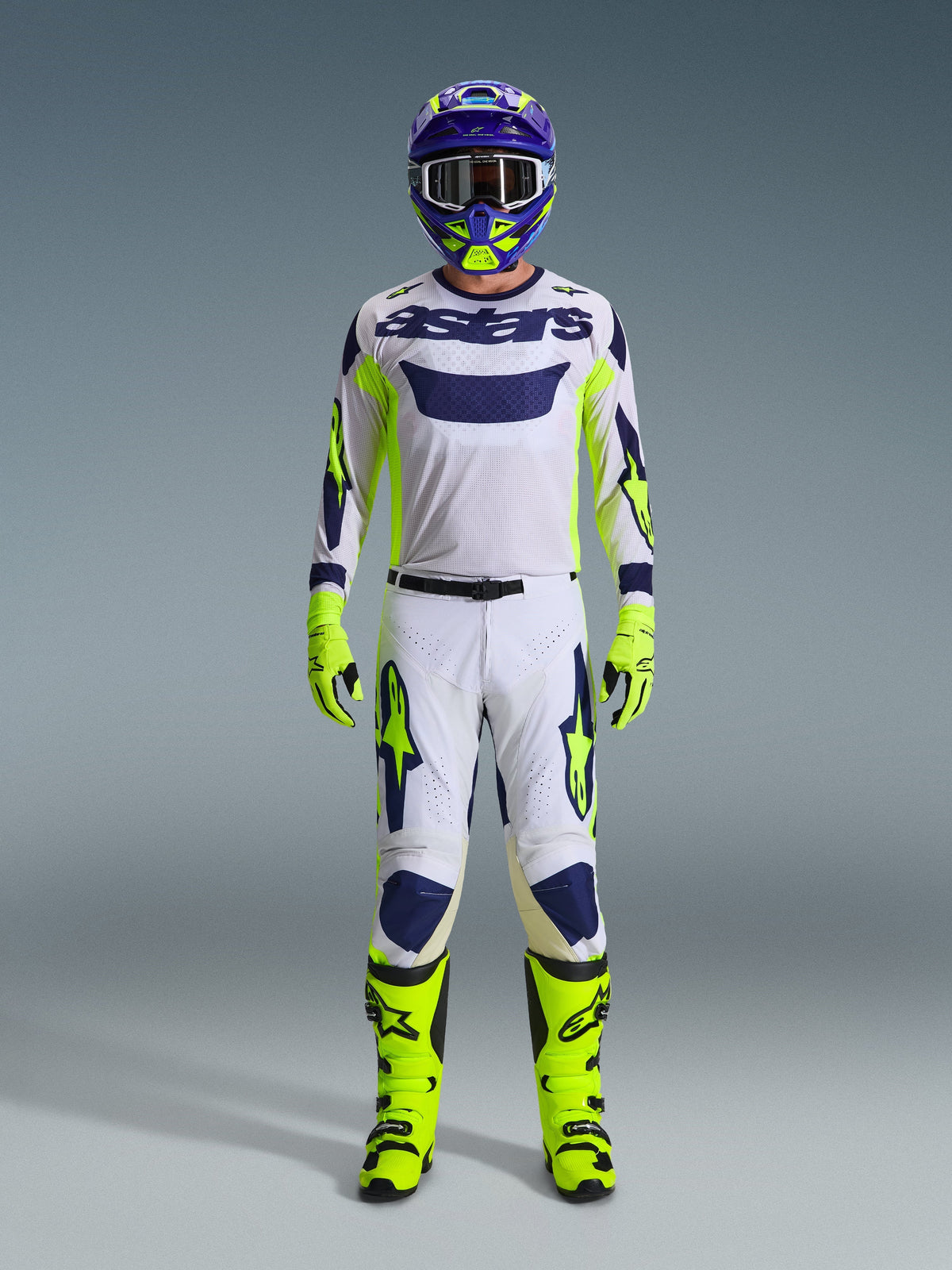 Motocyklista motocrossowy Alpinestars w pełnym stroju wyścigowym, jasnoszara koszulka Racer Air Riway z neonowymi żółtymi i niebieskimi akcentami, pasujące biało-żółte spodnie, neonowe żółte buty i rękawice, fioletowo-żółty kask, profesjonalny strój do wyścigów off-road