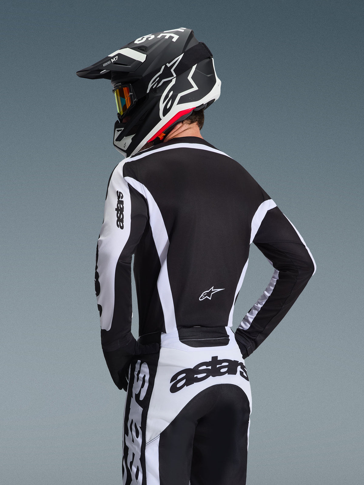 Zawodnik motocrossowy widoczny od tyłu, ubrany w koszulkę Alpinestars 2026 Racer Riway w kolorze biało-czarnym z logo „astars” na rękawach i w dolnej części pleców, w zestawie z dopasowanymi spodniami motocrossowymi oraz czarnym kaskiem z goglami, skoordynowany strój do wyścigów terenowych.