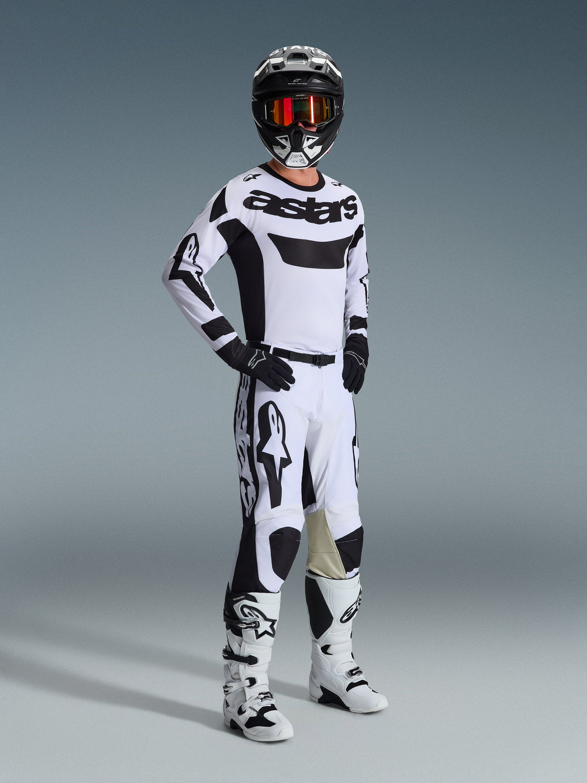 Motocyklista motocrossowy w pełnym stroju wyścigowym, koszulka Alpinestars 2026 Racer Riway w kolorze biało-czarnym, pasujące biało-czarne spodnie motocrossowe, czarny kask z lustrzanymi goglami, biało-czarne buty i czarne rękawice, skoordynowany zestaw do wyścigów terenowych