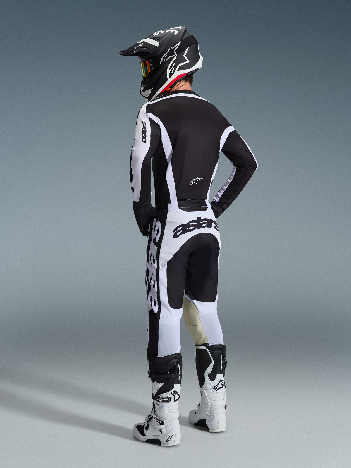 Kierowca motocrossowy w pełnym stroju wyścigowym, widok z tyłu, biało-czarna koszulka Alpinestars 2026 Racer Riway, dopasowane biało-czarne spodnie z wyraźnym brandingiem „astars”, białe buty i czarny kask z białymi akcentami, skoordynowany zestaw wyczynowy do wyścigów terenowych.