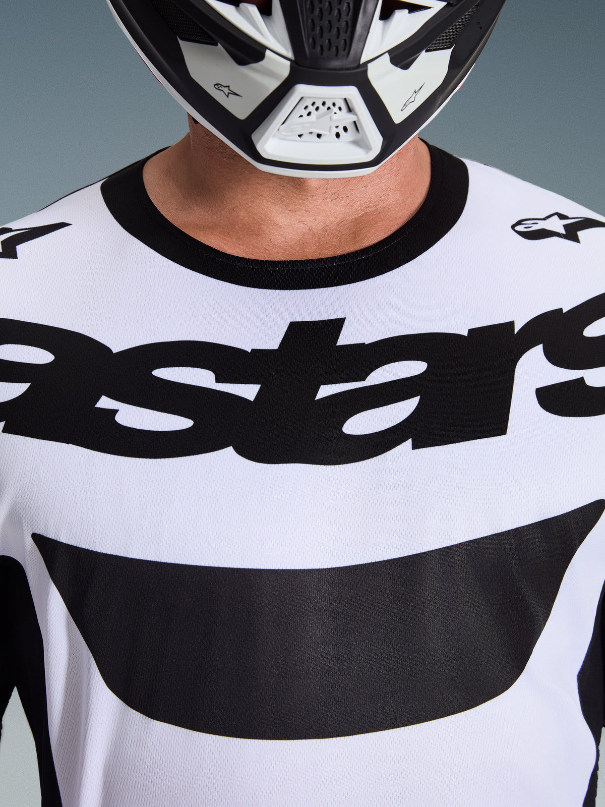 Alpinestars 2026 Racer Riway Jersey, koszulka motocrossowa, biało-czarna, zbliżenie na klatkę piersiową z wyraźnym czarnym logo astars i czarnym kołnierzykiem, widoczna w zestawie z biało-czarnym kaskiem, profesjonalna odzież do jazdy off-road