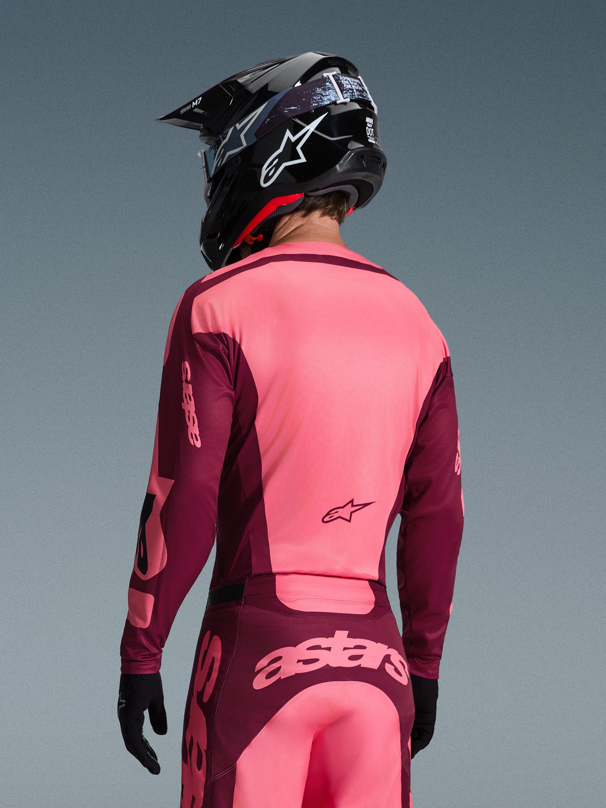 Motocross rider seen from behind wearing the Alpinestars 2026 Racer Riway Gear Set, Bright Pink and Maroon jersey and pants, black motocross helmet with white logo, and black gloves, featuring 'astars' branding on the back.