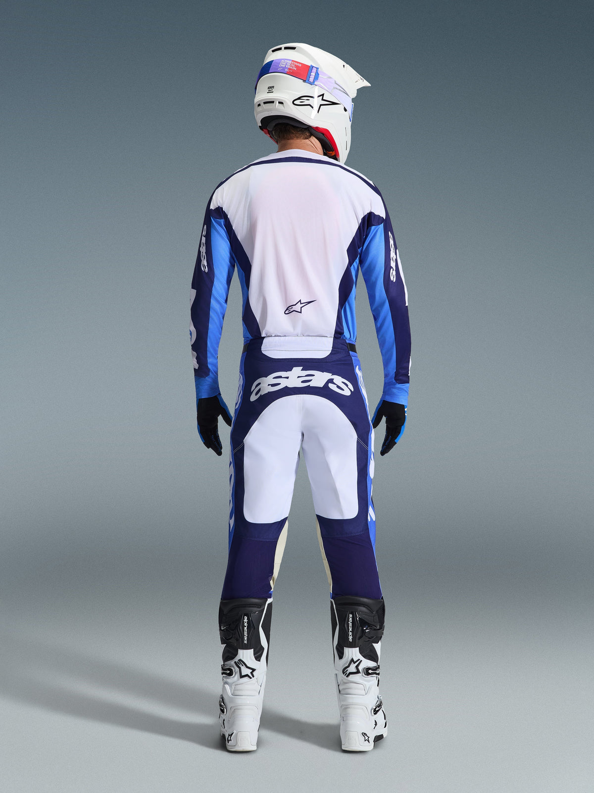 Zawodnik motocrossowy widziany od tyłu w koszulce Alpinestars 2026 Racer Riway w kolorze Ucla Blue i białym, pasujące spodnie motocrossowe z białymi i granatowymi panelami oraz logo Astars, biało-czarne buty i biały kask, kompletny, dopasowany strój wyścigowy do jazdy w terenie.