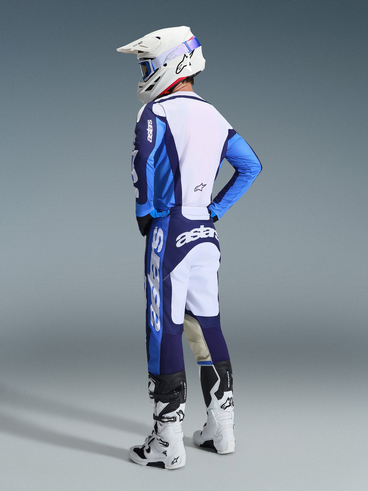 Motocross rider from a rear-side view wearing the Alpinestars 2026 Racer Riway Gear Set in UCLA Blue and White, featuring a blue and white jersey with 'astars' branding, matching blue and white pants, a white motocross helmet with blue goggles, and white and black Tech series boots.