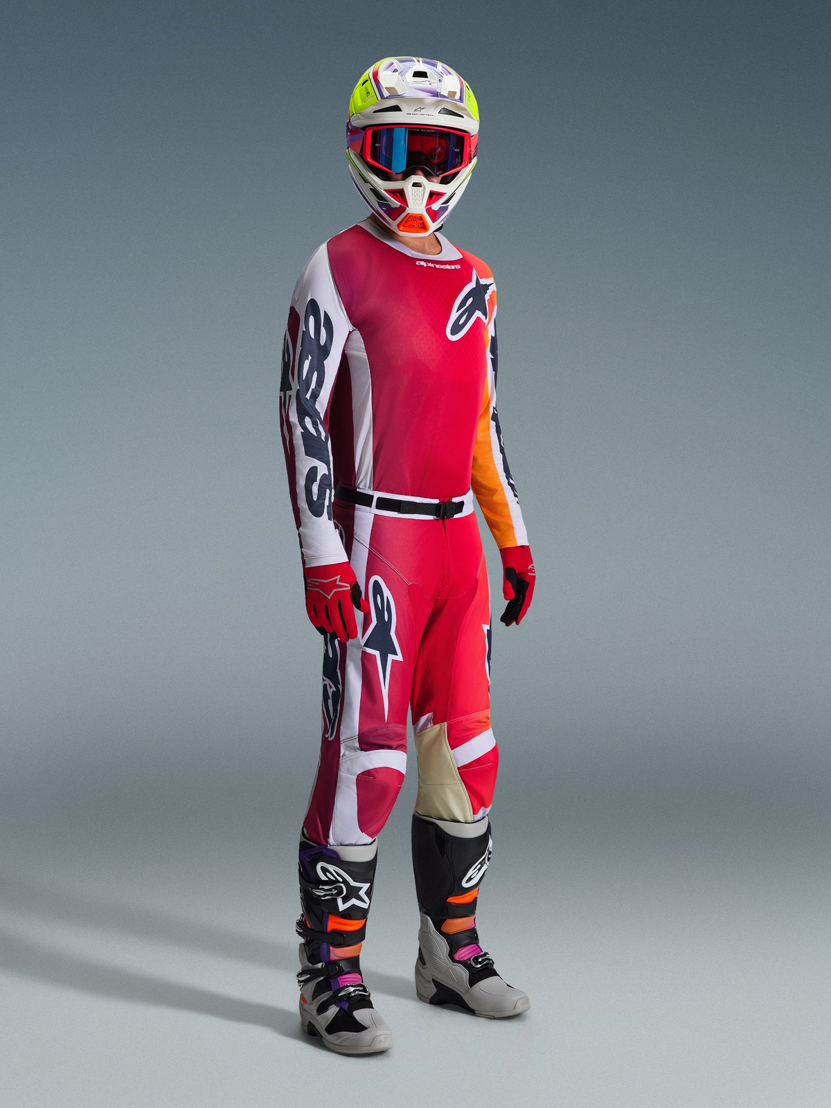 Motocross rider wearing complete racing gear, Alpinestars 2026 Racer Portl Jersey in red and light gray, matching motocross pants with white and dark green accents, multi-colored helmet with red goggles, gray and black boots with orange and purple details, red gloves, coordinated outfit for off-road racing