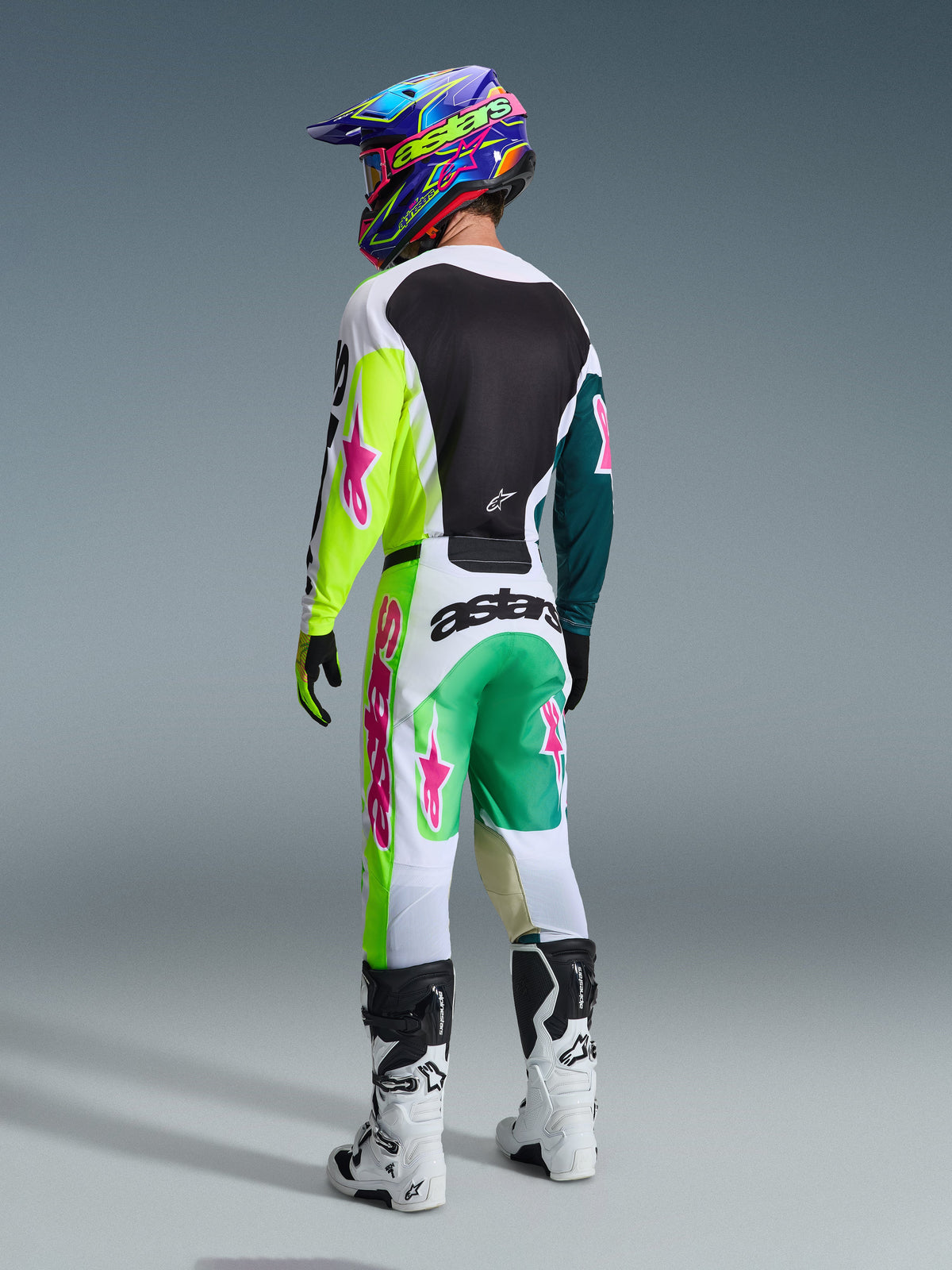 Motocyklista motocrossowy widziany od tyłu, zestaw Alpinestars 2026 Racer Portl, koszulka z neonowożółtymi i morskimi rękawami oraz różowymi logotypami, biało-zielone spodnie z różowymi akcentami, kolorowy kask i białe buty, profesjonalna odzież off-road