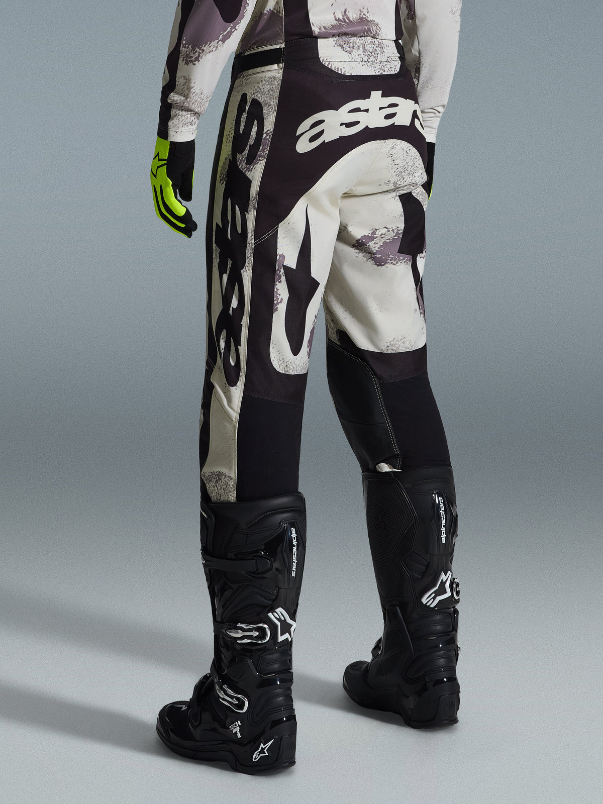 Alpinestars 2026 Racer Lahnd Pants, spodnie motocrossowe, kolor Sand Camo i szary, widok z tyłu z wyraźnym białym brandingiem na siedzeniu i nogawkach, w zestawieniu z czarnymi butami motocrossowymi i żółtymi rękawicami, profesjonalny sprzęt off-road