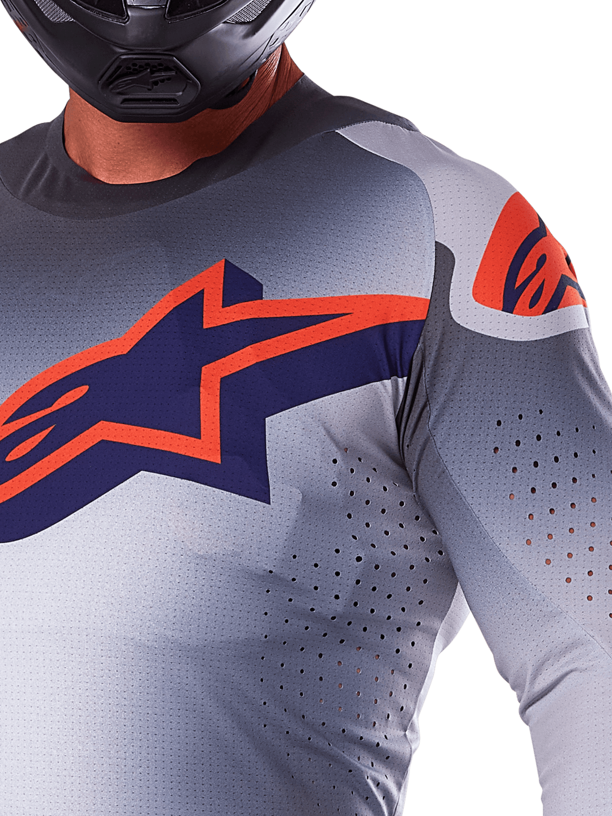 Zbliżenie na zawodnika w koszulce Alpinestars 2025 Supertech Lipan, koszulka motocrossowa, szara i pomarańczowa fluo, z dużym pomarańczowym logo Alpinestars na klatce piersiowej, laserowo wycinanymi otworami wentylacyjnymi i czarnym kaskiem, profesjonalny sprzęt wyścigowy.