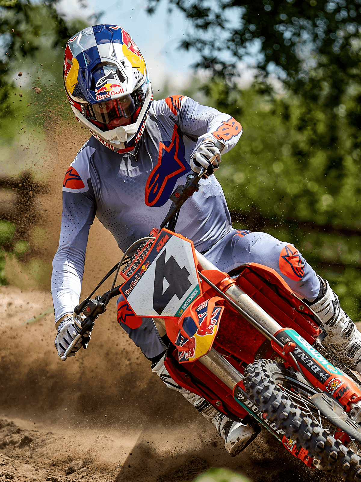 Motocrossista w akcji na torze ziemnym, koszulka Alpinestars 2025 Supertech Lipan w kolorze Gray Orange Fluo, pasujące szare spodnie z pomarańczowymi akcentami, białe buty i kask z logo Red Bull, pokonywanie zakrętu na pomarańczowym motocyklu crossowym z rozbryzgami ziemi, profesjonalny sprzęt wyścigowy off-road