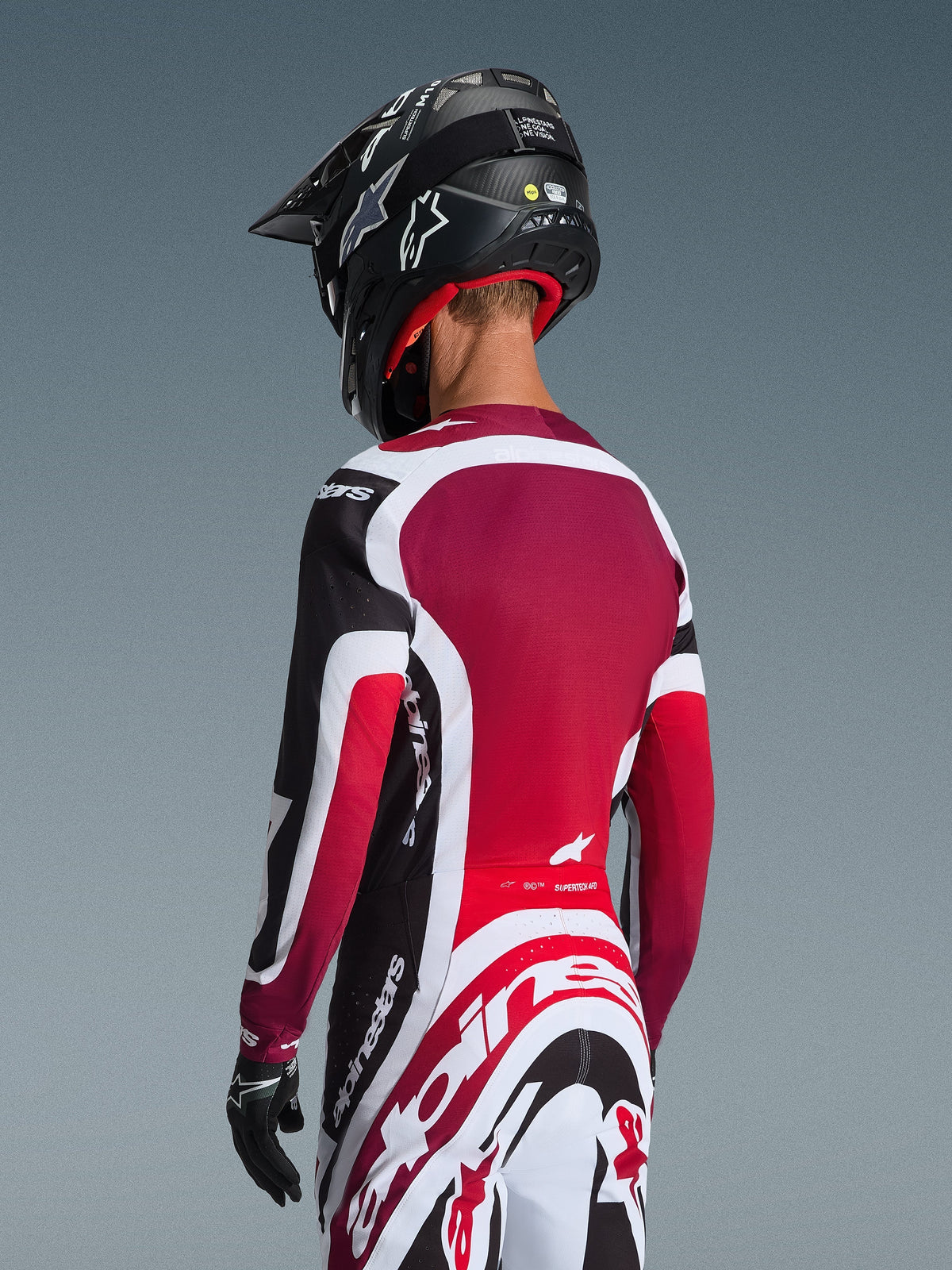 Widok z tyłu motocyklisty motocrossowego ubranego w koszulkę 2026 Supertech AFD Vista w kolorze czarno-czerwono-białym, zestawioną z dopasowanymi spodniami i czarnym kaskiem, z wyraźnym brandingiem Alpinestars, czerwonymi panelami z siatki i sportowym krojem wyścigowym.