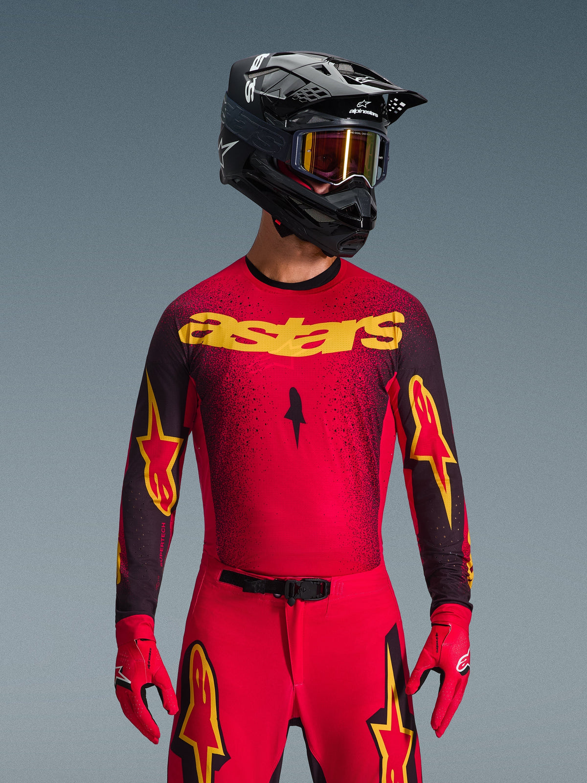 Koszulka Alpinestars 2026 Supertech Scenz, koszulka motocrossowa, czerwona, czarna i żółta, noszona przez motocyklistę w dopasowanym czarnym kasku i czerwonych rękawicach, z wyraźnym żółtym logo „astars” na klatce piersiowej i czarnym gradientowym nakrapianym wzorem na ramionach.