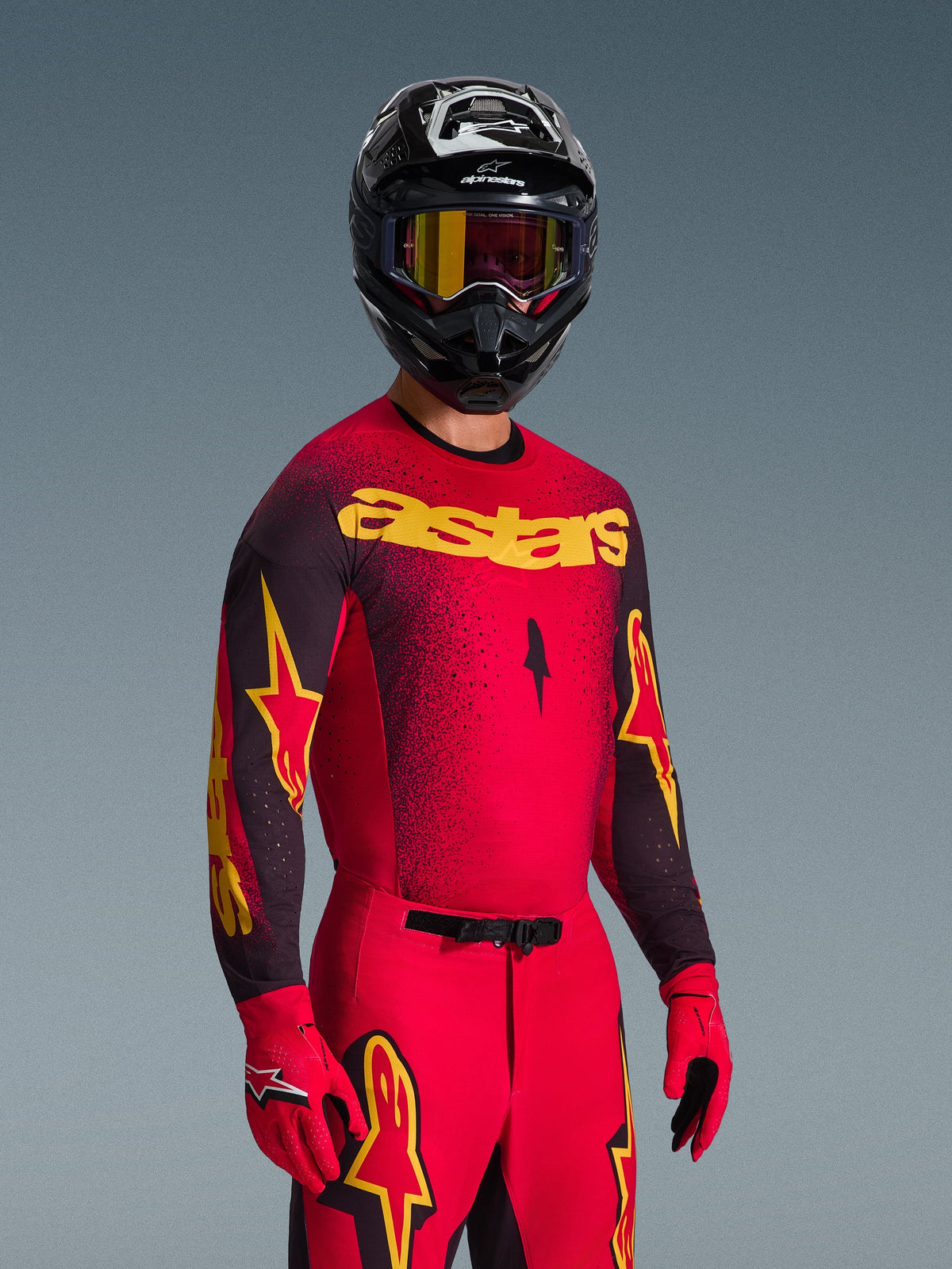 Zawodnik motocrossowy w koszulce Alpinestars 2026 Supertech Scenz, czerwono-czarna z żółtym brandingiem „astars”, pasujące czerwone spodnie i rękawice, czarny kask z goglami, profesjonalny zestaw odzieży wyścigowej do motocrossu