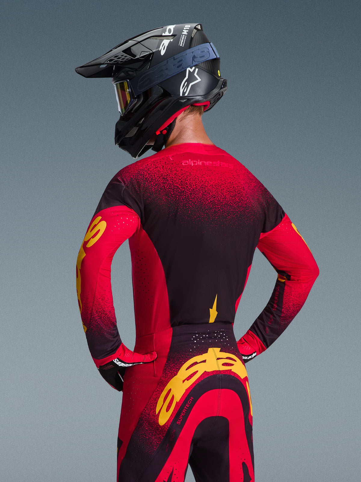 Alpinestars Supertech Scenz Jersey 2026, czerwono-czarno-żółta koszulka motocrossowa, widok z tyłu na zawodnika w dopasowanym stroju wyścigowym, spodniach off-road i czarnym kasku, czerwono-czarny gradient i wyraziste żółte logo Alpinestars