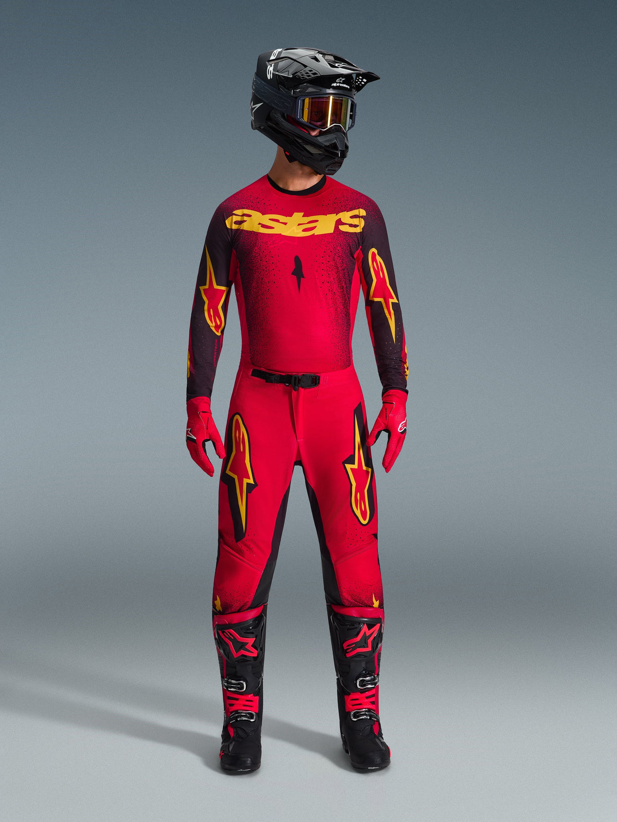Zawodnik motocrossowy w koszulce Alpinestars 2026 Supertech Scenz w kolorze czerwono-czarno-żółtym, w zestawie z dopasowanymi spodniami, czarnym kaskiem z goglami, czerwonymi rękawicami oraz czarno-czerwonymi butami, profesjonalny zestaw odzieży wyścigowej do jazdy off-road