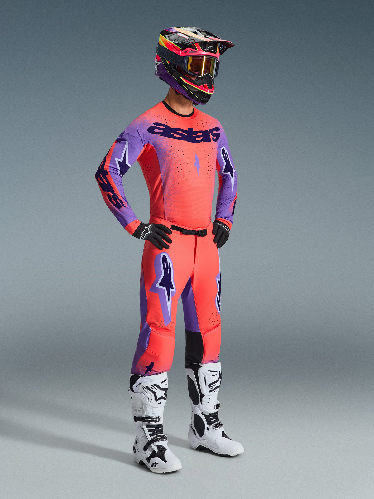 Zawodnik motocrossowy ubrany w kompletny zestaw Alpinestars 2026 Supertech Scenz, koszulka w kolorze Hot Coral z fioletowymi rękawami i logo 'astars', koralowe spodnie z fioletowymi akcentami, wielokolorowy kask ze złotymi goglami, biało-czarne buty i czarne rękawice, profesjonalny strój wyścigowy do jazdy w terenie.