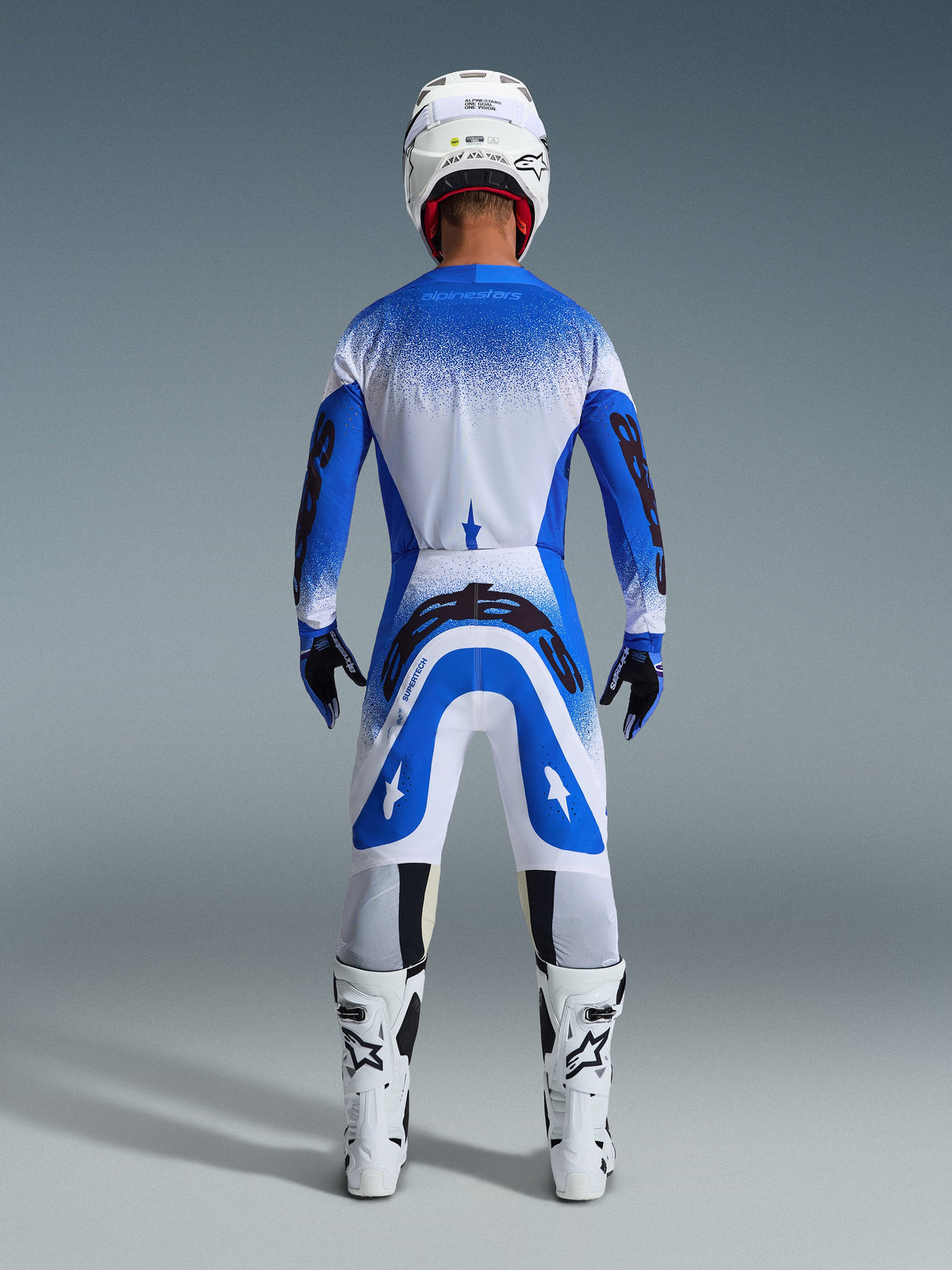 Zawodnik motocrossowy widziany z tyłu w zestawie Alpinestars 2026 Supertech Scenz w kolorze Ucla Blue White, niebiesko-biała koszulka z czarnym brandingiem, pasujące spodnie, biały kask i białe buty, profesjonalny sprzęt wyścigowy do motocrossu off-road