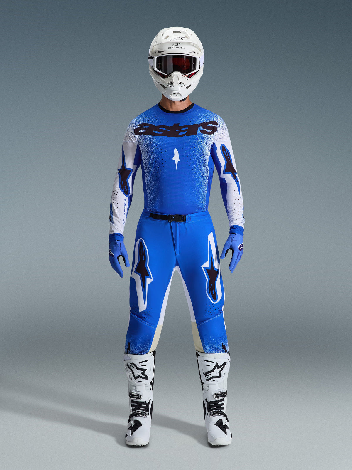 Zawodnik motocrossowy ubrany w zestaw Alpinestars 2026 Supertech Scenz w kolorystyce Ucla Blue i białej, zawierający lekką niebiesko-białą koszulkę z czarnym brandingiem, dopasowane niebieskie spodnie, biały kask wyścigowy i białe buty motocrossowe, profesjonalna odzież wyczynowa do wyścigów terenowych.