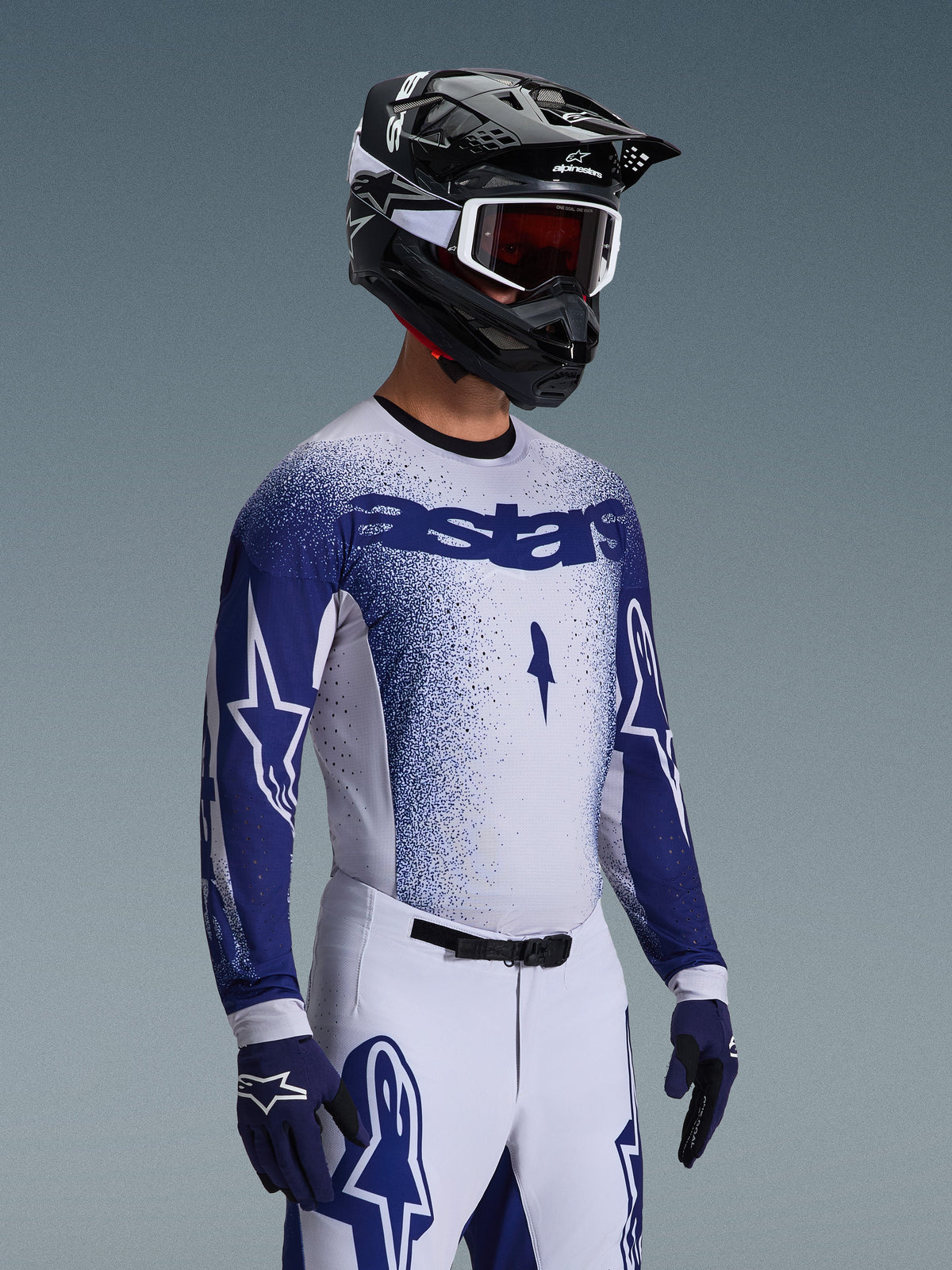 Zawodnik motocrossowy w koszulce 2026 Supertech Scenz w kolorze jasnoszarym i ciemnoniebieskim, w połączeniu z pasującymi biało-niebieskimi spodniami wyścigowymi, niebieskimi rękawicami oraz czarnym kaskiem z białymi goglami. Koszulka charakteryzuje się niebieskim efektem gradientu kropkowego na klatce piersiowej i rękawach, profesjonalnym sportowym krojem i wyraźnym brandingiem 'astars'.