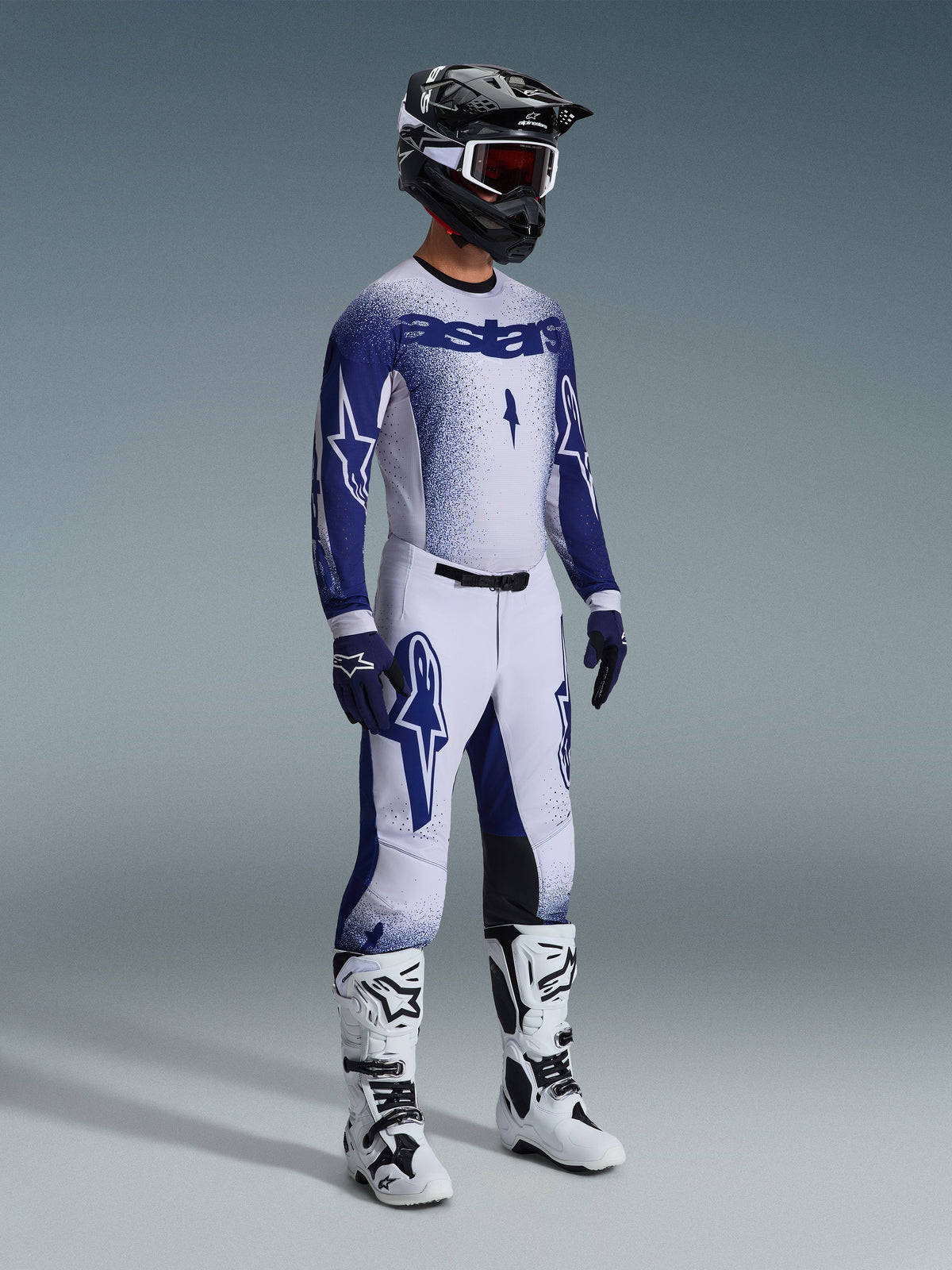 Motocyklista motocrossowy w kompletnym profesjonalnym stroju wyścigowym, koszulka 2026 Supertech Scenz w kolorze jasnoszarym i ciemnoniebieskim, pasujące spodnie motocrossowe z grafiką gwiazdy, czarny kask z czerwonymi goglami, biało-czarne buty wyczynowe, dopasowane niebiesko-białe rękawice do wyścigów terenowych