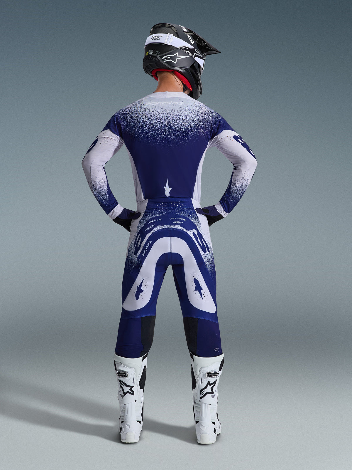 Motocrossista widziany od tyłu w koszulce Alpinestars 2026 Supertech Scenz, jasnoszaro-ciemnoniebieskiej z kropkowanym wzorem gradientowym, pasujące spodnie motocrossowe, białe buty wyścigowe i czarny kask, kompletny strój wyścigowy off-road