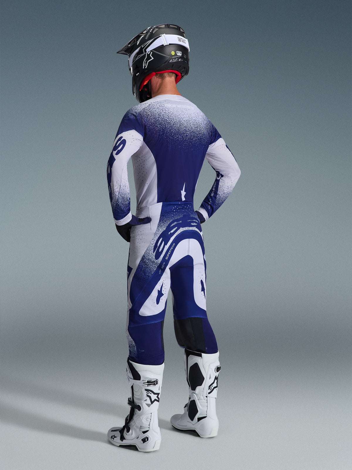 Widok z tyłu zawodnika motocrossowego w koszulce Alpinestars 2026 Supertech Scenz w kolorze jasnoszarym i ciemnoniebieskim, kropkowany wzór gradientowy, pasujące spodnie i białe buty motocrossowe, profesjonalny sportowy strój wyścigowy