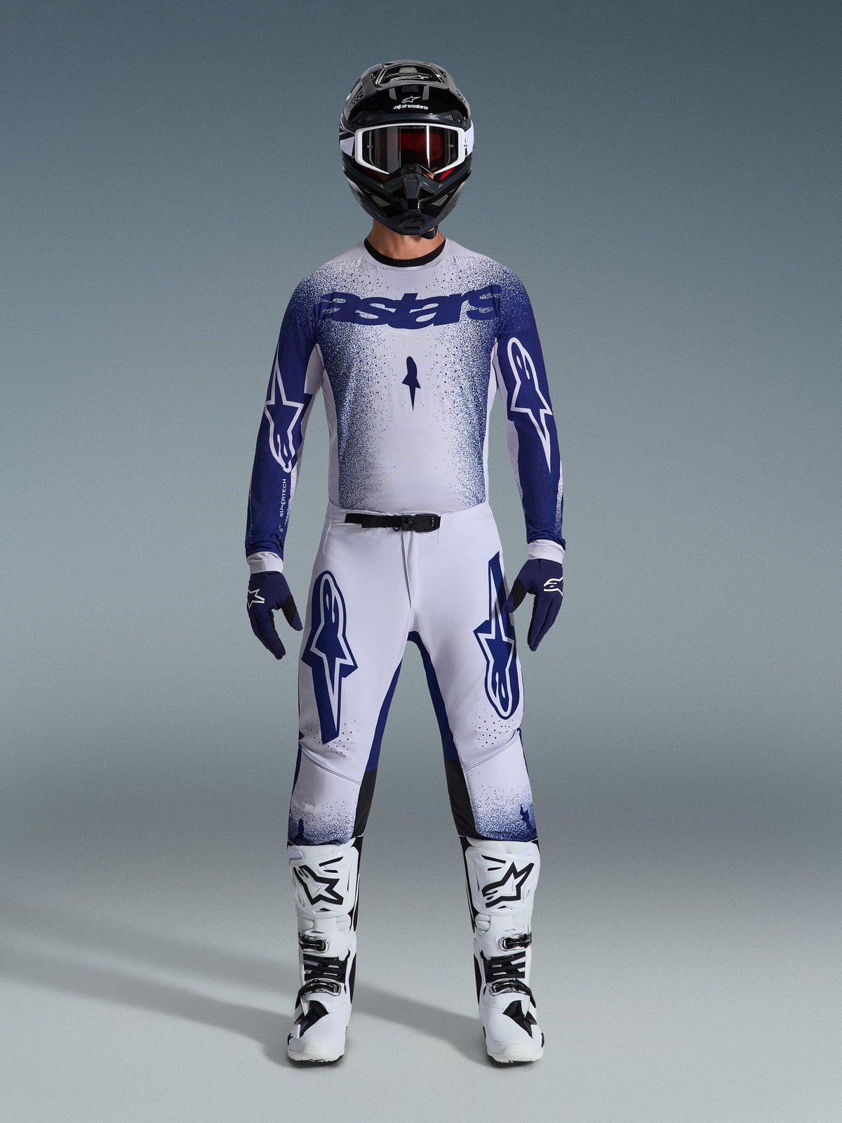 Zawodnik motocrossowy Alpinestars w pełnym zestawie wyścigowym, koszulka 2026 Supertech Scenz w jasnoszarym i ciemnoniebieskim kolorze, pasujące jasnoszare spodnie z niebieskim logo, czarno-biały kask z czerwonymi goglami, biało-czarne buty, ciemnoniebieskie rękawice, skoordynowany zestaw do wyścigów off-road