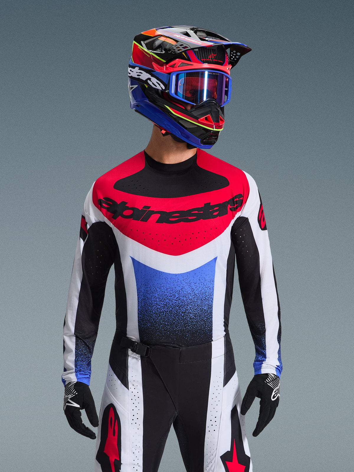 Alpinestars 2026 Techstar Knif Gear Set, zawodnik motocrossowy w pełnym stroju wyścigowym, czerwono-biało-niebieska koszulka z dużym czarnym brandingiem, pasujące czarno-białe spodnie z czerwonym logo gwiazdy, wielokolorowy kask z niebieskimi goglami i czarnymi rękawicami, skoordynowany zestaw do zawodów off-road