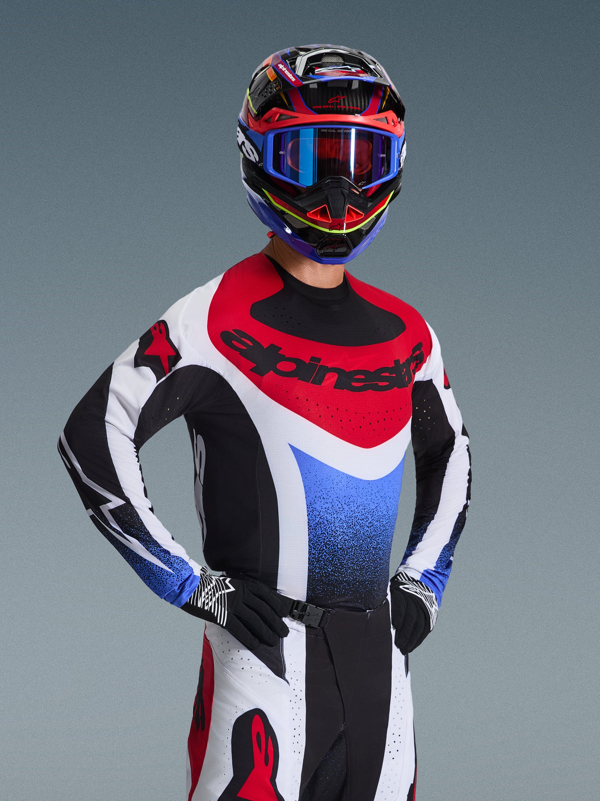 Zestaw Alpinestars Techstar Knif 2026, zawodnik motocrossowy w czerwono-biało-niebieskiej koszulce wyścigowej z czarnymi akcentami, dopasowany wielokolorowy kask i gogle, czarne rękawice, profesjonalny sprzęt na zawody motocrossowe.