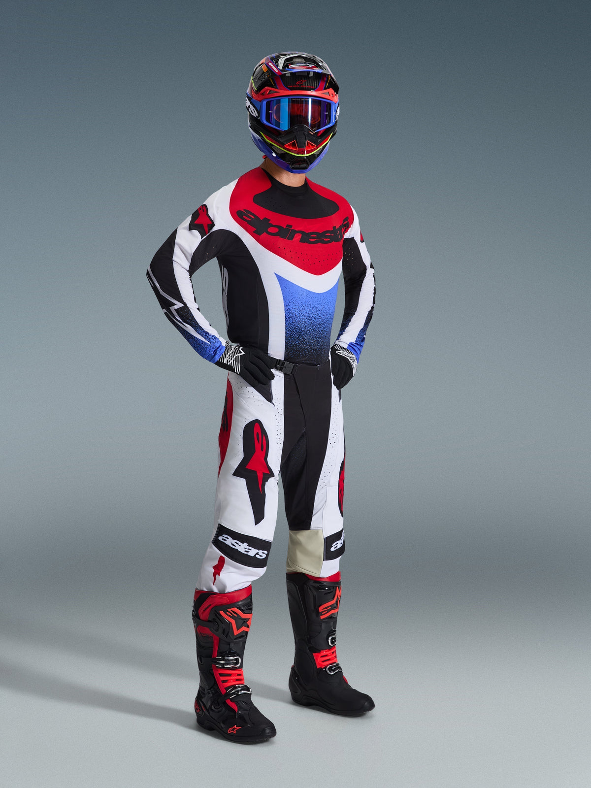 Kierowca motocrossowy w zestawie Alpinestars 2026 Techstar Knif, koszulka z czarnymi, białymi i czerwonymi panelami oraz niebieskim gradientem na klatce piersiowej, białe spodnie z czerwonymi logotypami Alpinestars i czarnymi akcentami, czarno-czerwone buty, kompletny profesjonalny zestaw do wyścigów off-road