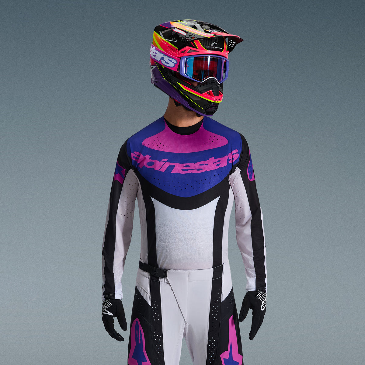 Zestaw Alpinestars Techstar Knif 2026, zawodnik motocrossowy w bluzie z niebiesko-różową grafiką i białym korpusem, białe spodnie z czarnymi panelami i różowym logo, fioletowo-różowy kask z dopasowanymi goglami, czarne rękawice, profesjonalny strój do wyścigów terenowych