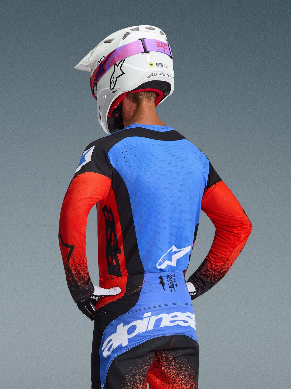 Widok od tyłu zawodnika motocrossowego w koszulce 2026 Techstar Knif w kolorystyce Orange Black UCLA Blue, z niebieskim panelem tylnym z laserowo wyciętymi perforacjami, pomarańczowymi rękawami i czarnymi akcentami, w zestawie z pasującymi spodniami motocrossowymi i białym kaskiem Alpinestars.