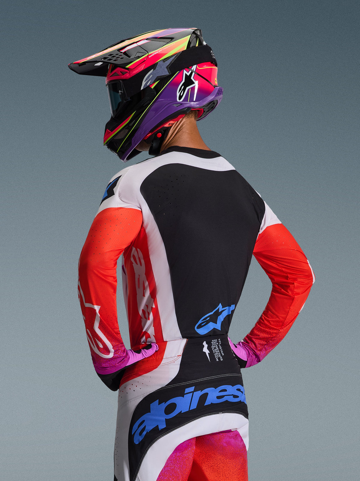 Motocyklista motocrossowy od tyłu w zestawie Alpinestars 2026 Techstar Knif, jaskrawopomarańczowe rękawy z czarnym panelem na plecach, biało-czarne spodnie z niebieskim logo, intensywnie fioletowo-różowy kask, profesjonalna odzież motocrossowa do jazdy w terenie