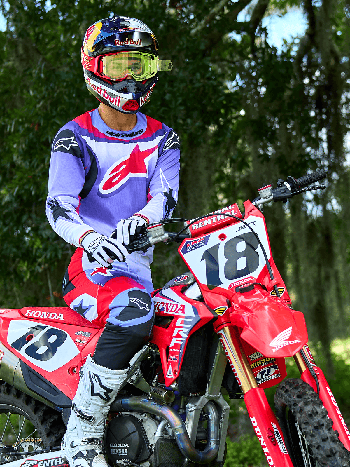 Zawodnik motocrossowy na czerwonym motocyklu Honda CRF, koszulka Alpinestars 2025 Fluid Wurx fioletowo-jaskrawoczerwona, pasujące fioletowo-czerwone spodnie, kask Red Bull z żółtymi goglami, białe buty i rękawice Alpinestars, profesjonalny sprzęt MX do jazdy terenowej