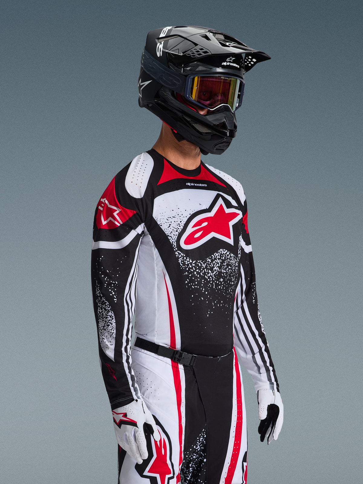 Zawodnik motocrossowy ubrany w koszulkę Alpinestars 2026 Techstar Nomur w kolorystyce Black White Red, z dużym czerwonym logo Astars i nakrapianymi wzorami graficznymi, w połączeniu z pasującymi spodniami wyścigowymi, białymi rękawicami i czarnym kaskiem z goglami, profesjonalny strój do rywalizacji off-road.