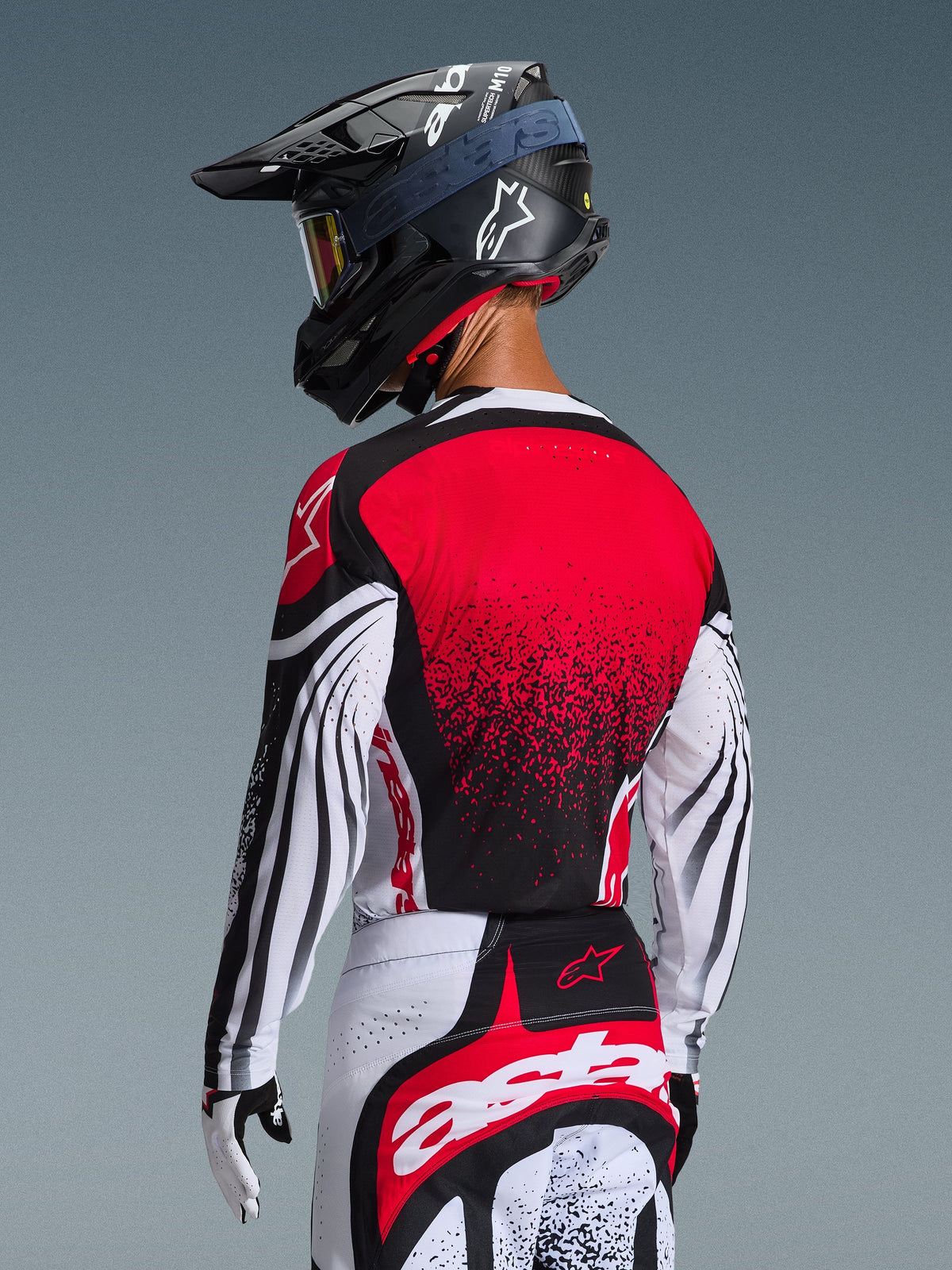Motocyklista motocrossowy widoczny od tyłu, koszulka Alpinestars 2026 Techstar Nomur, czerwony tył z czarnym wzorem gradientowym, białe rękawy z czarnymi akcentami, dopasowane czerwono-białe spodnie, czarny kask i białe rękawice, profesjonalny strój wyścigowy off-road