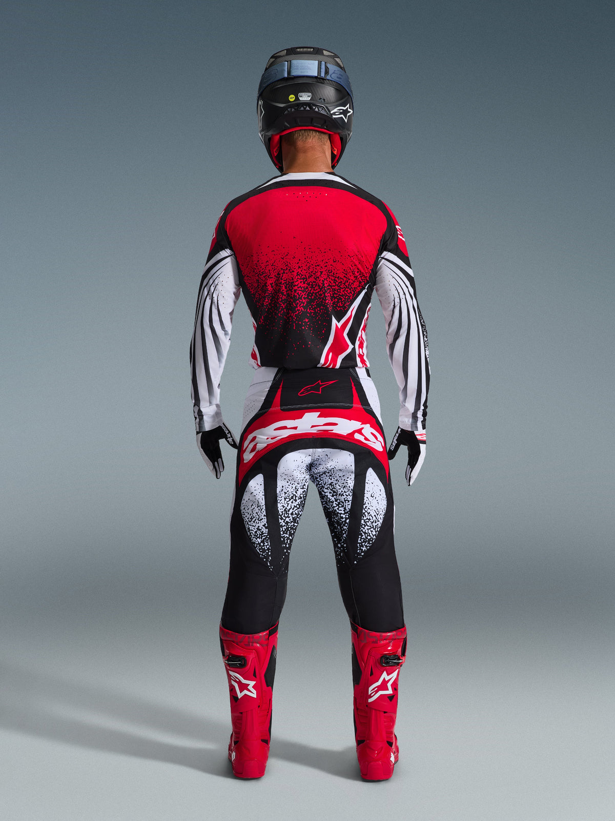 Motocyklista motocrossowy widziany od tyłu w zestawie Alpinestars 2026 Techstar Nomur, czerwono-biała koszulka z czarnym nakrapianym gradientem, pasujące biało-czerwone spodnie z czarnymi akcentami, jaskrawoczerwone buty i czarny kask, dopasowany strój do wyścigów terenowych