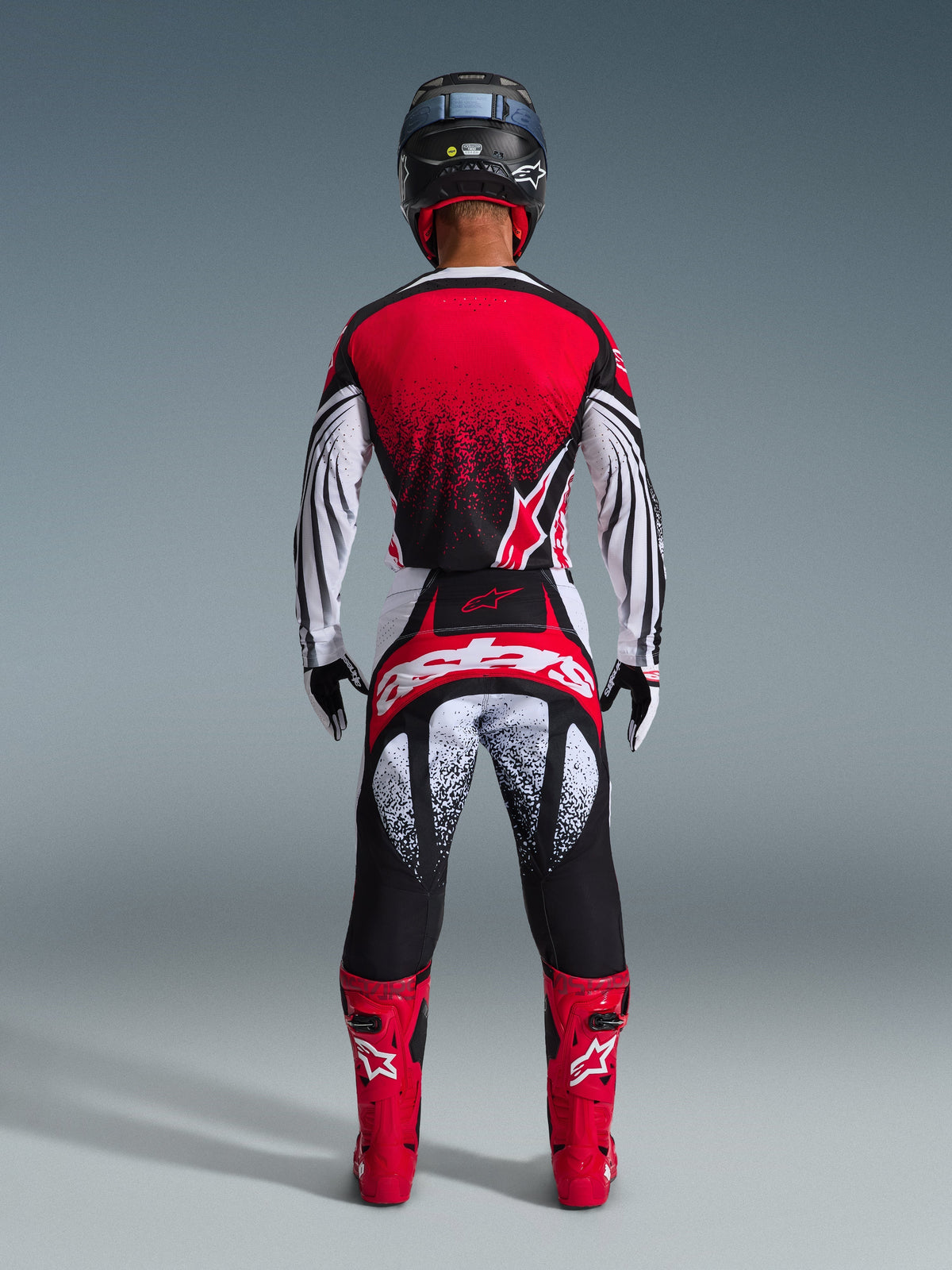 Kierowca motocrossowy widoczny od tyłu, ubrany w koszulkę Alpinestars 2026 Techstar Nomur, czerwono-czarno-biała z kropkowanym wzorem, pasujące spodnie wyścigowe, czerwone buty motocrossowe i czarny kask, kompletny zestaw odzieży do zawodów off-road