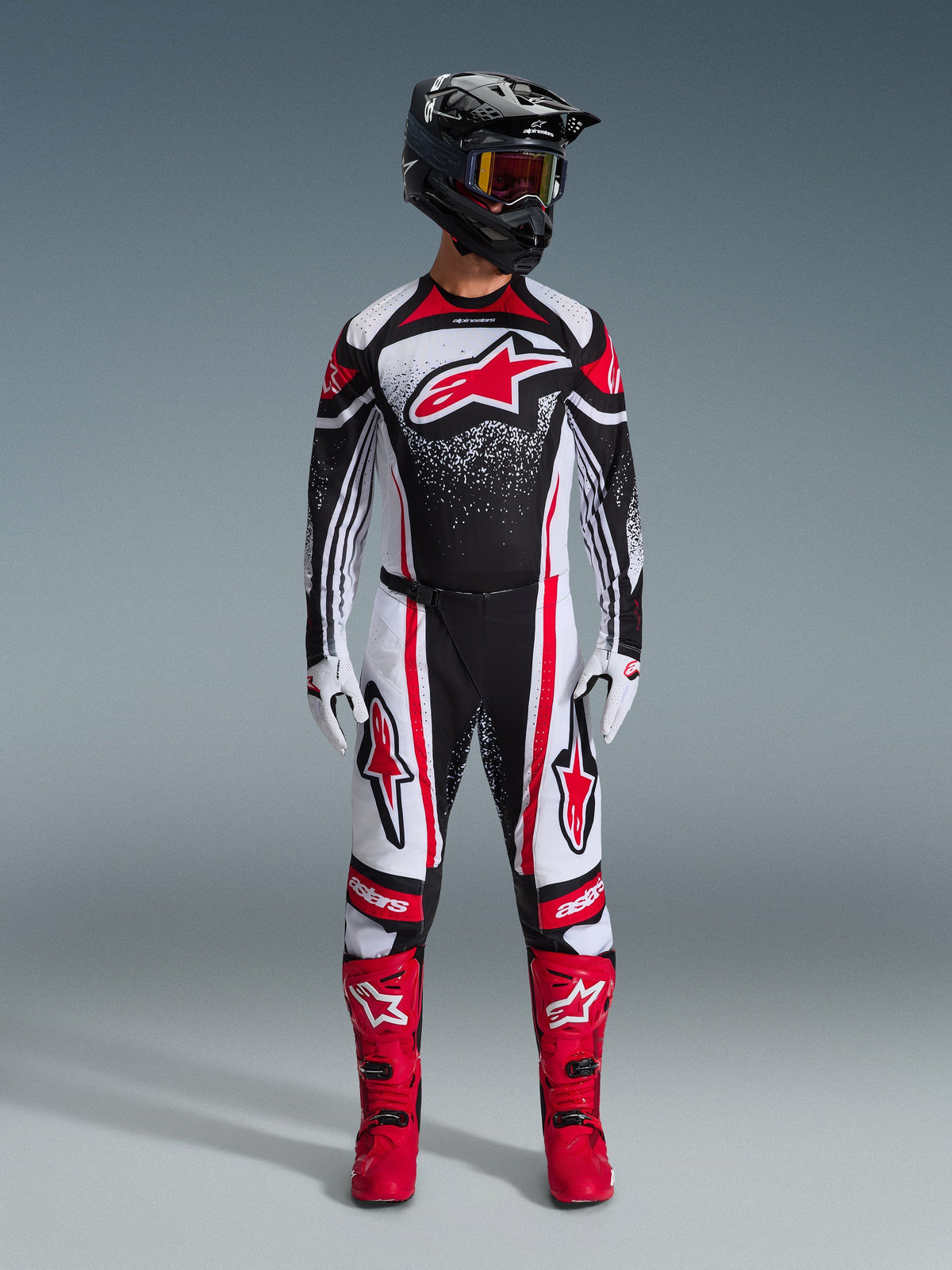 Motocyklista motocrossowy w pełnym stroju wyścigowym Alpinestars, koszulka 2026 Techstar Nomur w kolorze czarno-biało-czerwonym, pasujące spodnie z czerwonymi akcentami, czarny kask i jaskrawoczerwone buty, duże czerwone logo Astars na klatce piersiowej z nakrapianym wzorem gradientowym, dopasowany zestaw do zawodów off-road