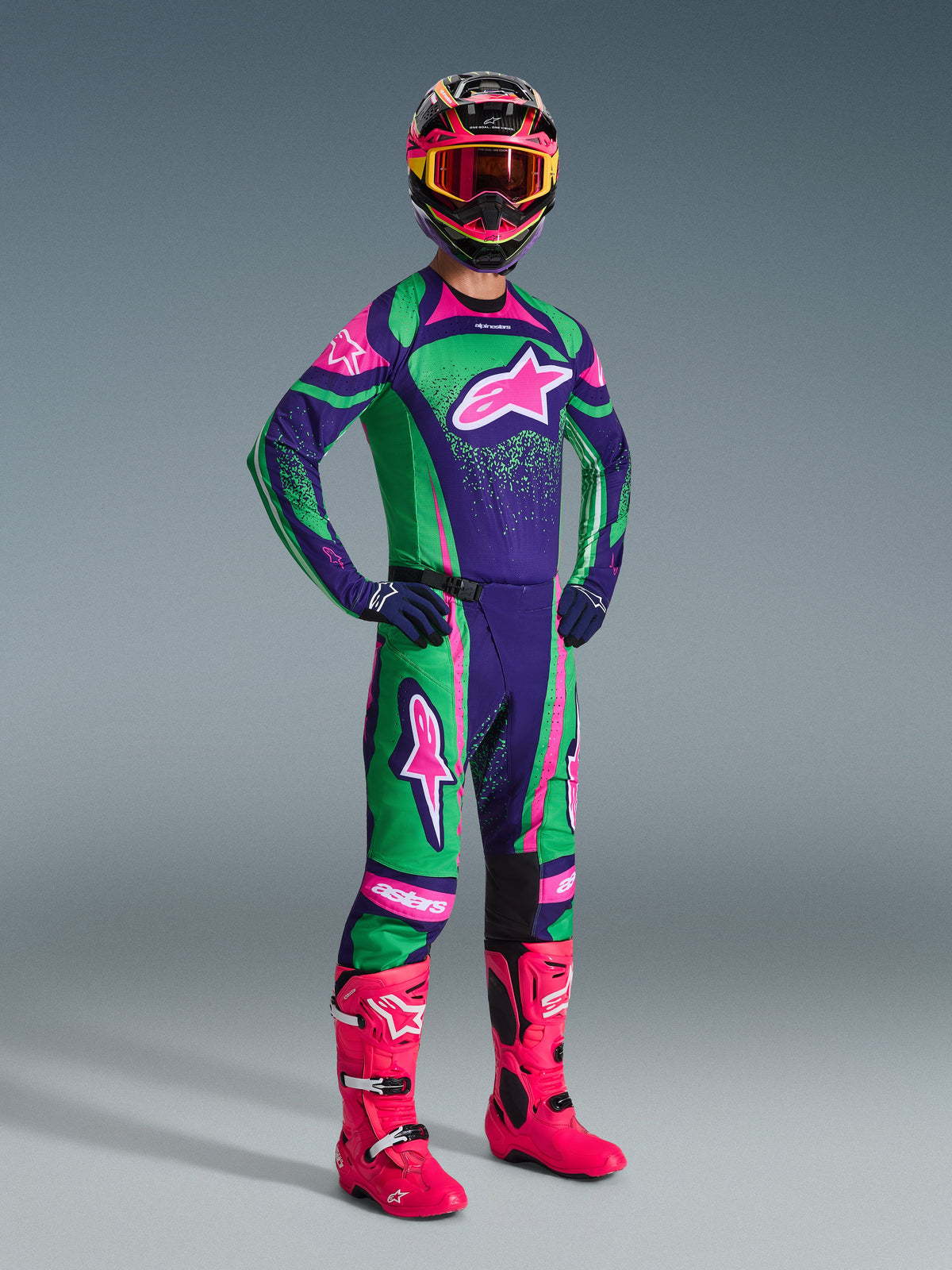 Motocyklista motocrossowy ubrany w zestaw Alpinestars 2026 Techstar Nomur w kolorystyce Deep Purple, Green i Pink Fluo, zawierający dopasowaną koszulkę i spodnie, kolorowy kask z różowymi goglami oraz jaskraworóżowe buty, profesjonalny sprzęt do wyścigów off-road.