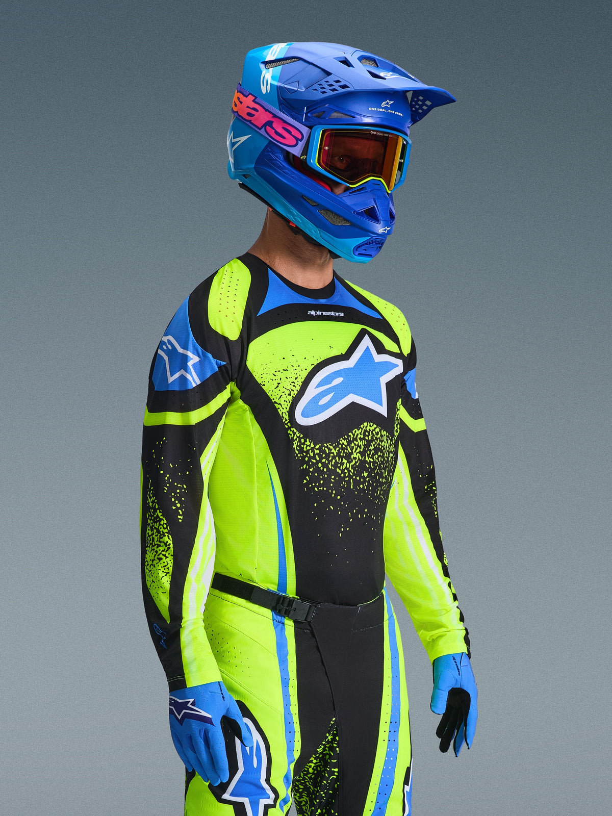 Zawodnik motocrossowy w koszulce Techstar Nomur 2026 w kolorze ciemnogranatowym i żółtym fluo, w zestawie z dopasowanymi spodniami, niebieskim kaskiem wyścigowym i goglami, z dużym logo Alpinestars i atletycznym krojem wyścigowym do jazdy terenowej.