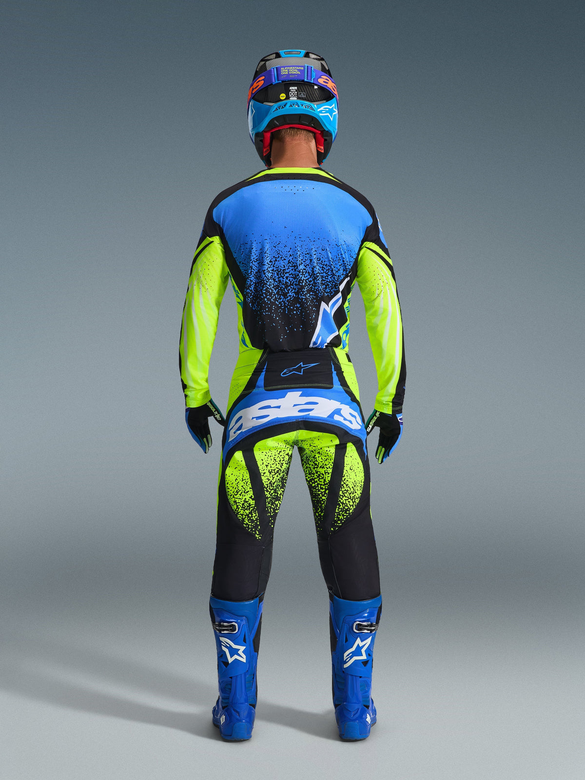 Widok od tyłu motocyklisty motocrossowego w koszulce 2026 Techstar Nomur w kolorach Dark Navy, Yellow Fluo i UCLA Blue, dopasowane żółto-czarne spodnie z brandingiem „Astars”, niebieskie buty wyścigowe i pasujący kask, kompletny profesjonalny strój do wyścigów terenowych.