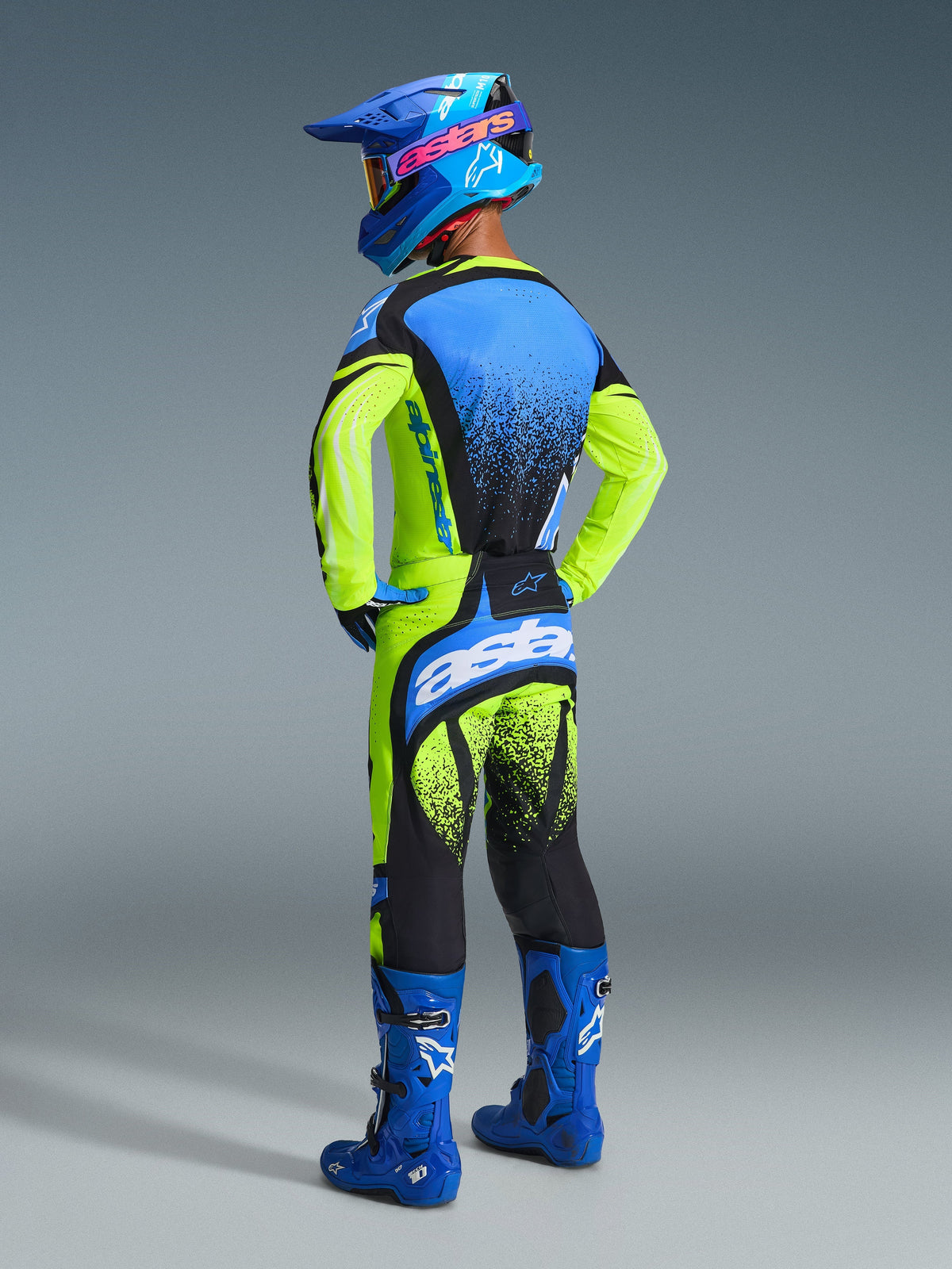 Motocyklista motocrossowy widoczny od tyłu w pełnym stroju wyścigowym, koszulka Alpinestars Techstar Nomur 2026 w kolorze żółtym fluo i niebieskim, pasujące żółto-czarne spodnie z niebieskimi akcentami, niebieski kask z różowym logo Alpinestars, niebieskie buty, ujęcie studyjne podkreślające tylny design i sportowy krój.